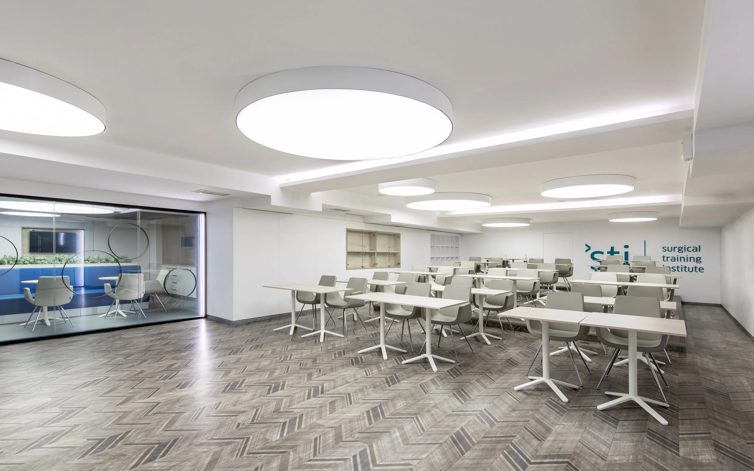 Empty training room with white tables, gray chairs, and bright circular ceiling lights, located at the surgical training institute.