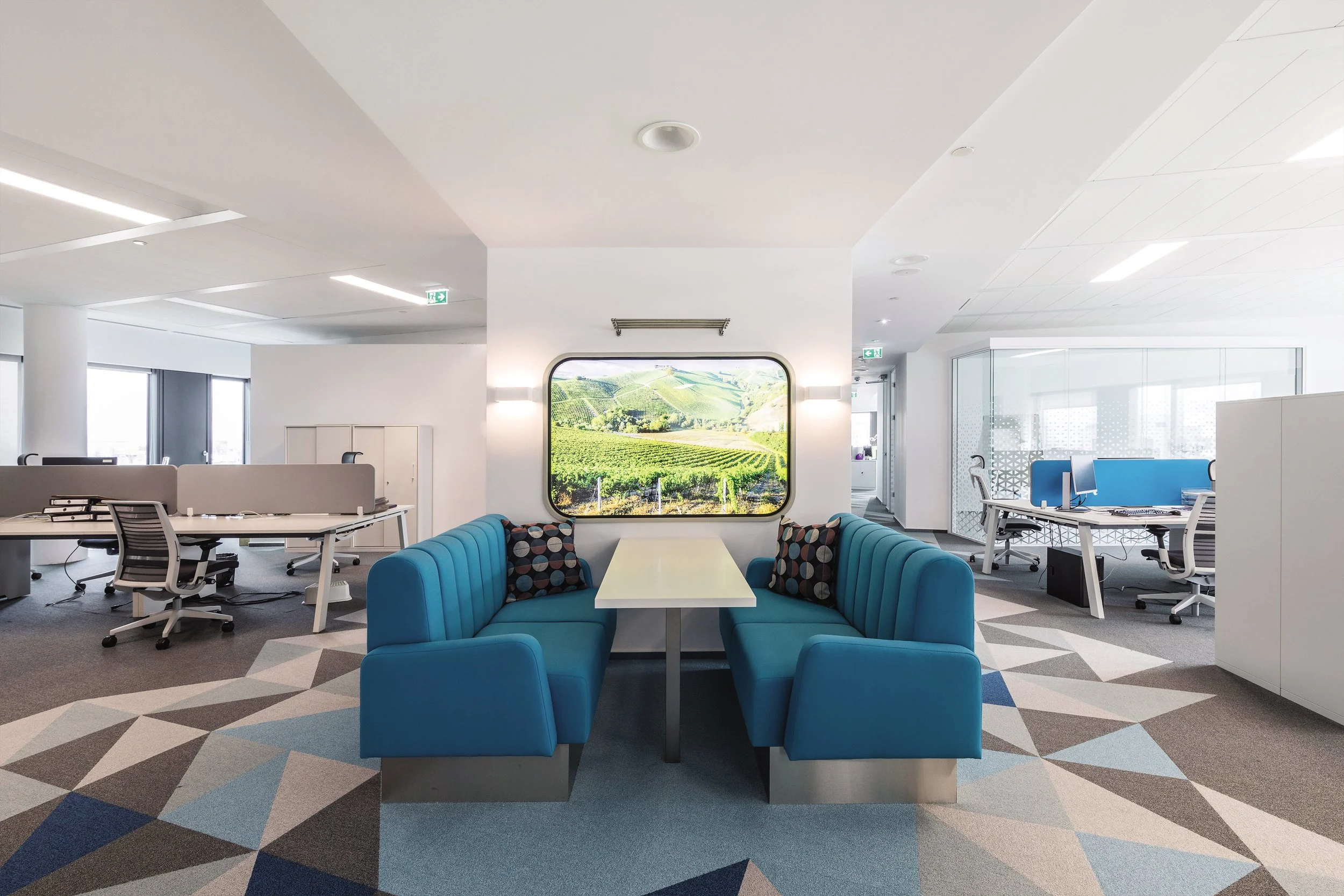 Modern office space with a blue booth seating area in the foreground, a scenic landscape picture on the wall, open workstations with chairs and computers on the left and right, and a bright, airy atmosphere.