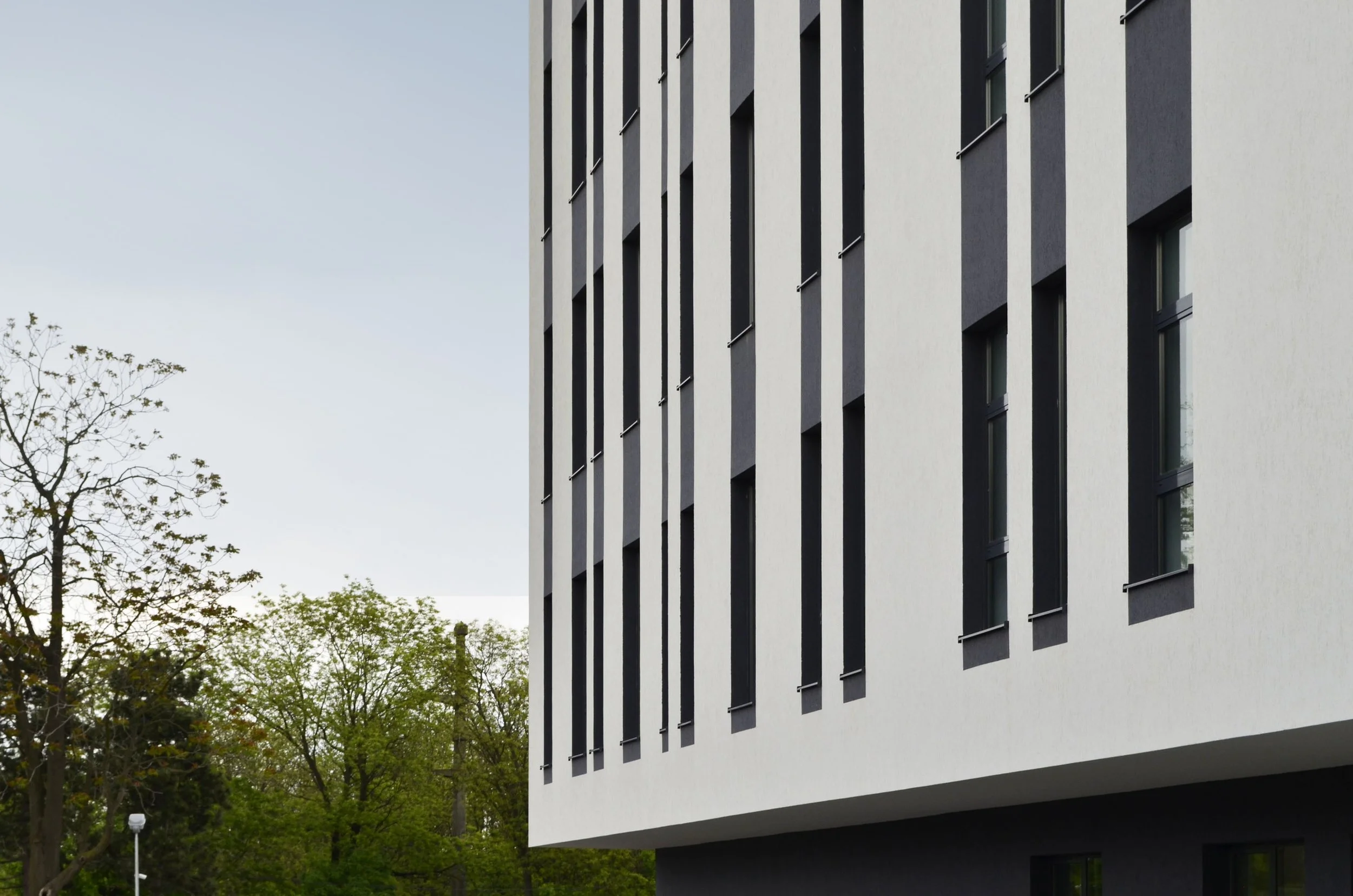 Modern building with narrow vertical black window frames on a white facade, trees with green foliage nearby.