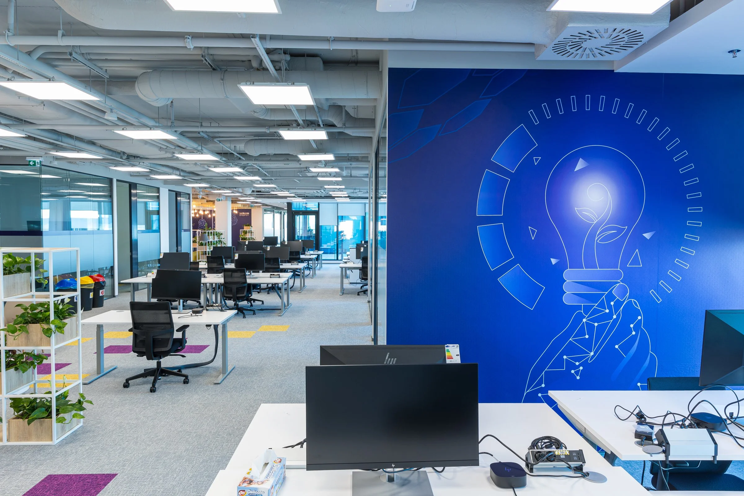Modern office workspace with desks, chairs, plants, and a blue wall featuring a lightbulb with a plant inside, symbolizing innovation and growth.