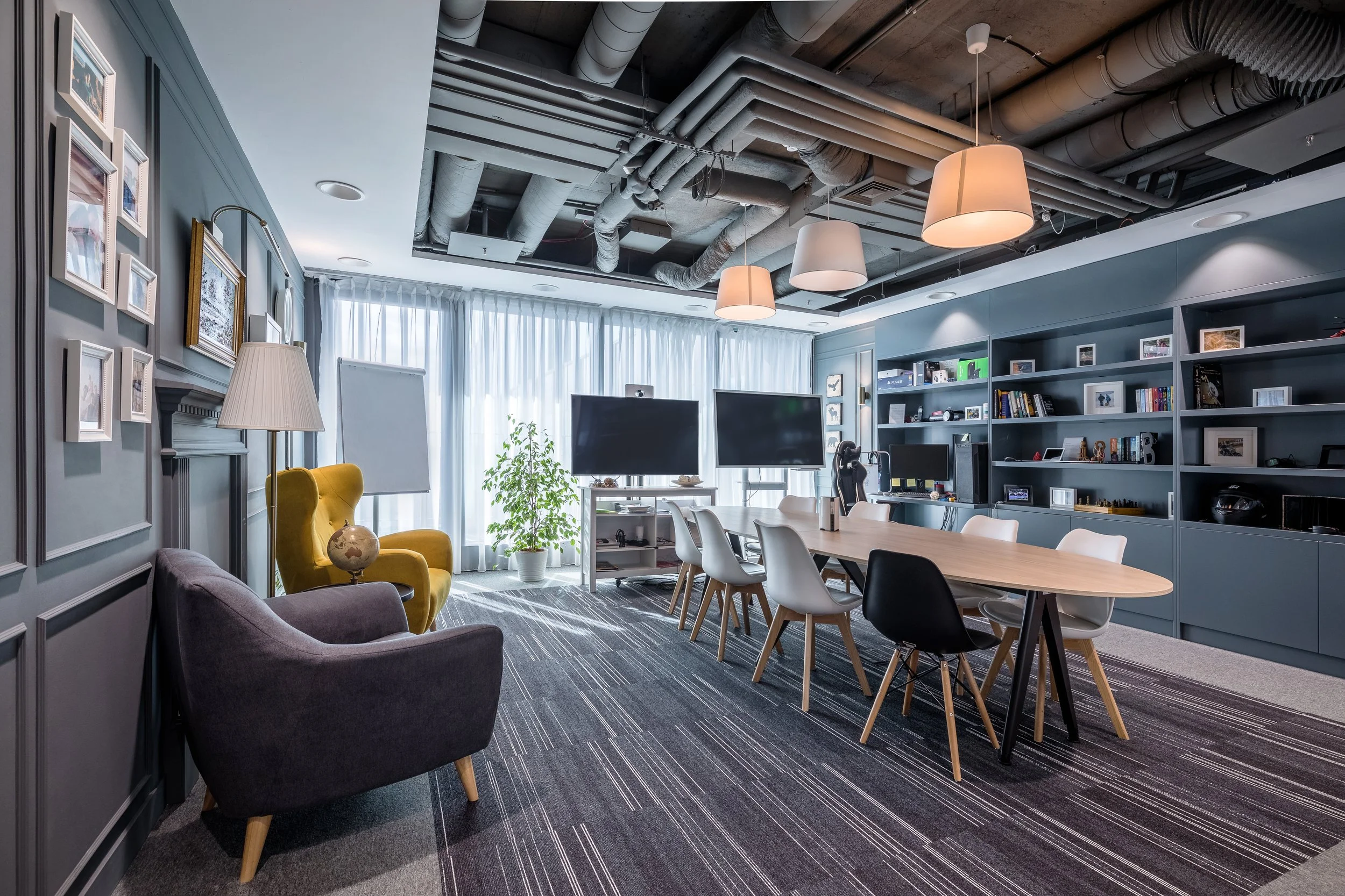 Modern office conference room with a large table, multiple chairs, a bookshelf filled with books and photos, two large monitors, armchairs, a floor lamp, a potted plant, and a ceiling with exposed ducts and hanging lamps.