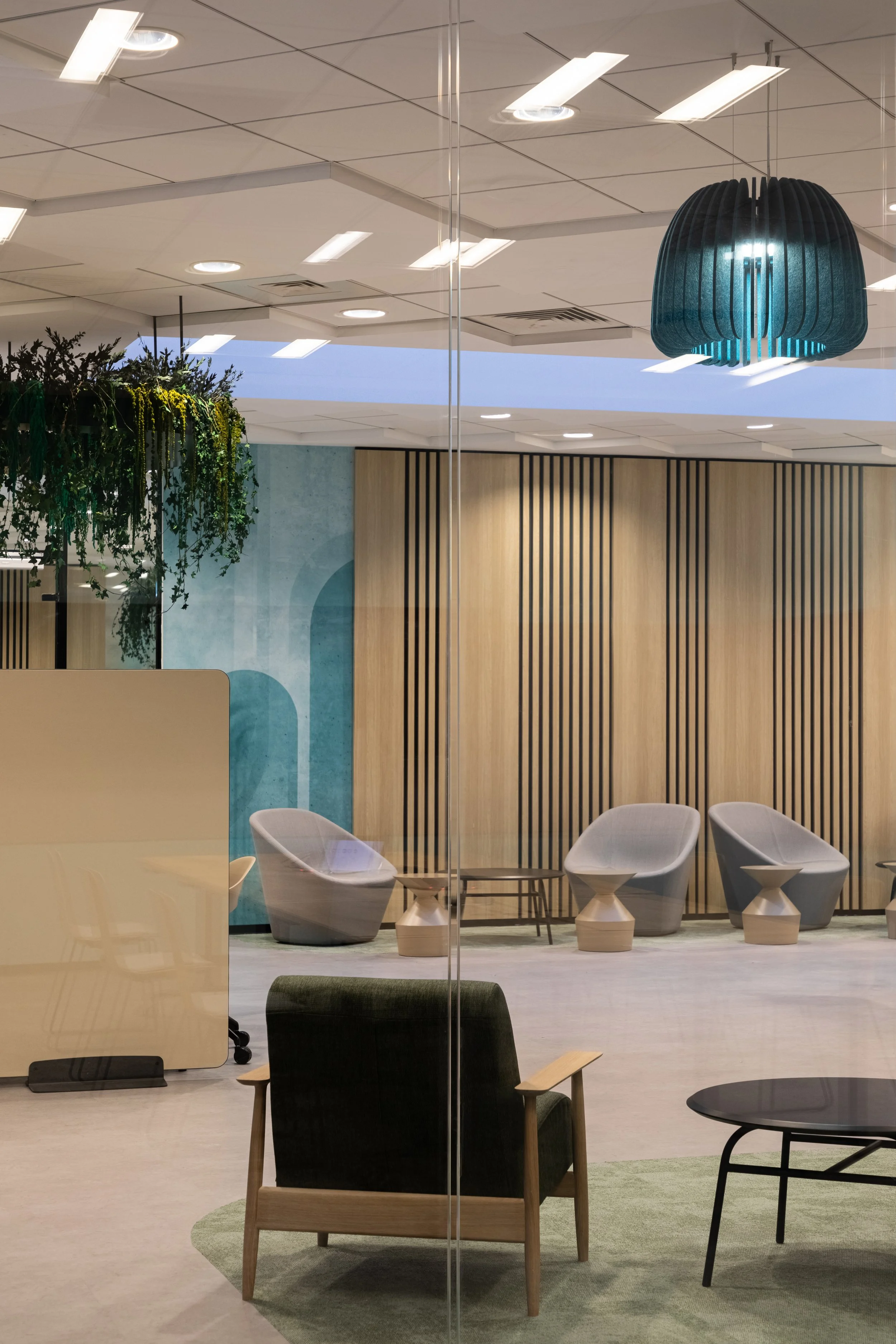 Modern office lounge with black armchair, round black table, and white chairs in the background, with wooden panel wall, green plants, and a teal pendant light overhead.