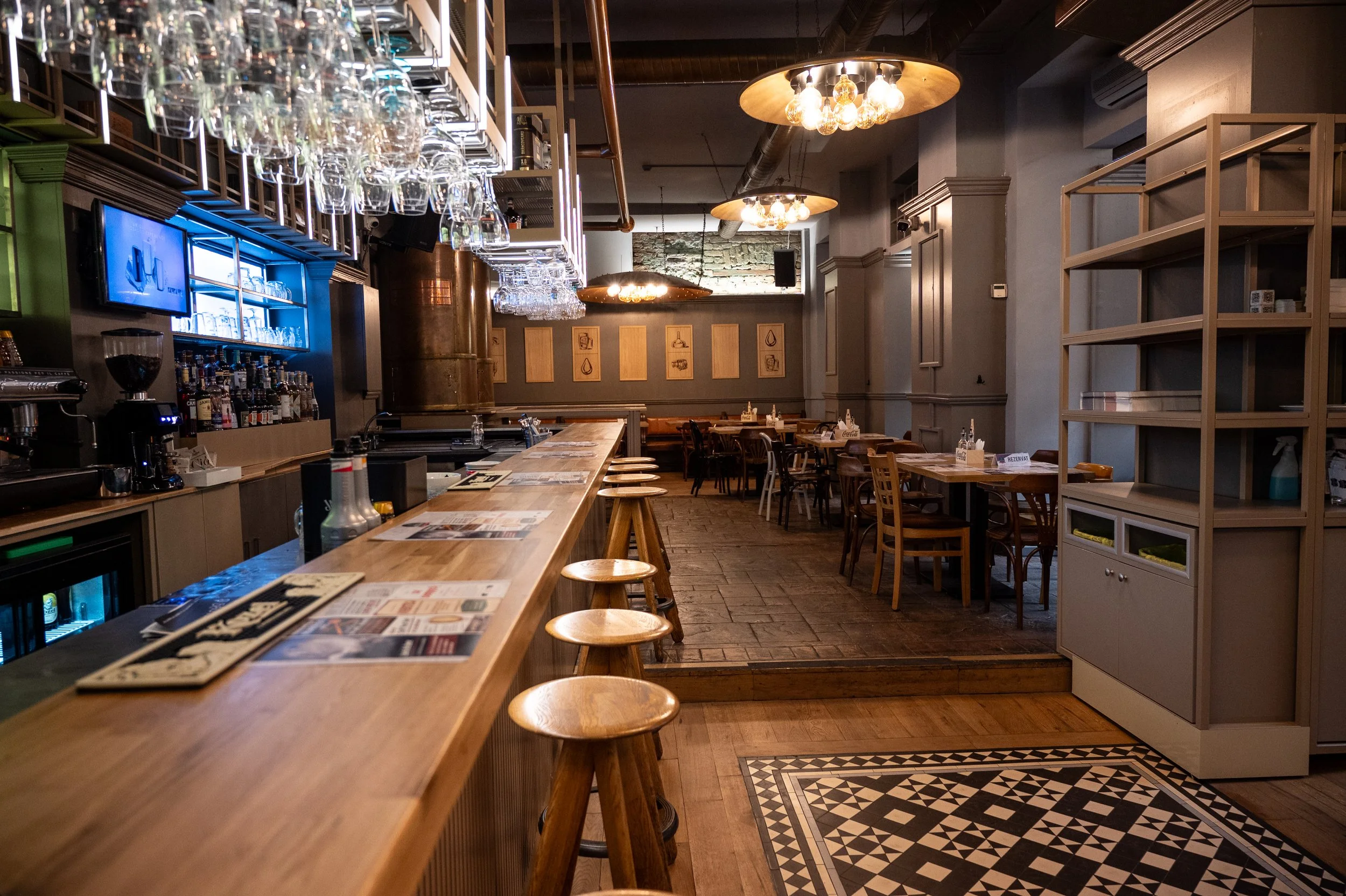 Interior of a restaurant with a wooden bar counter and stools, hanging glass racks, tables and chairs, wall art, and warm lighting.