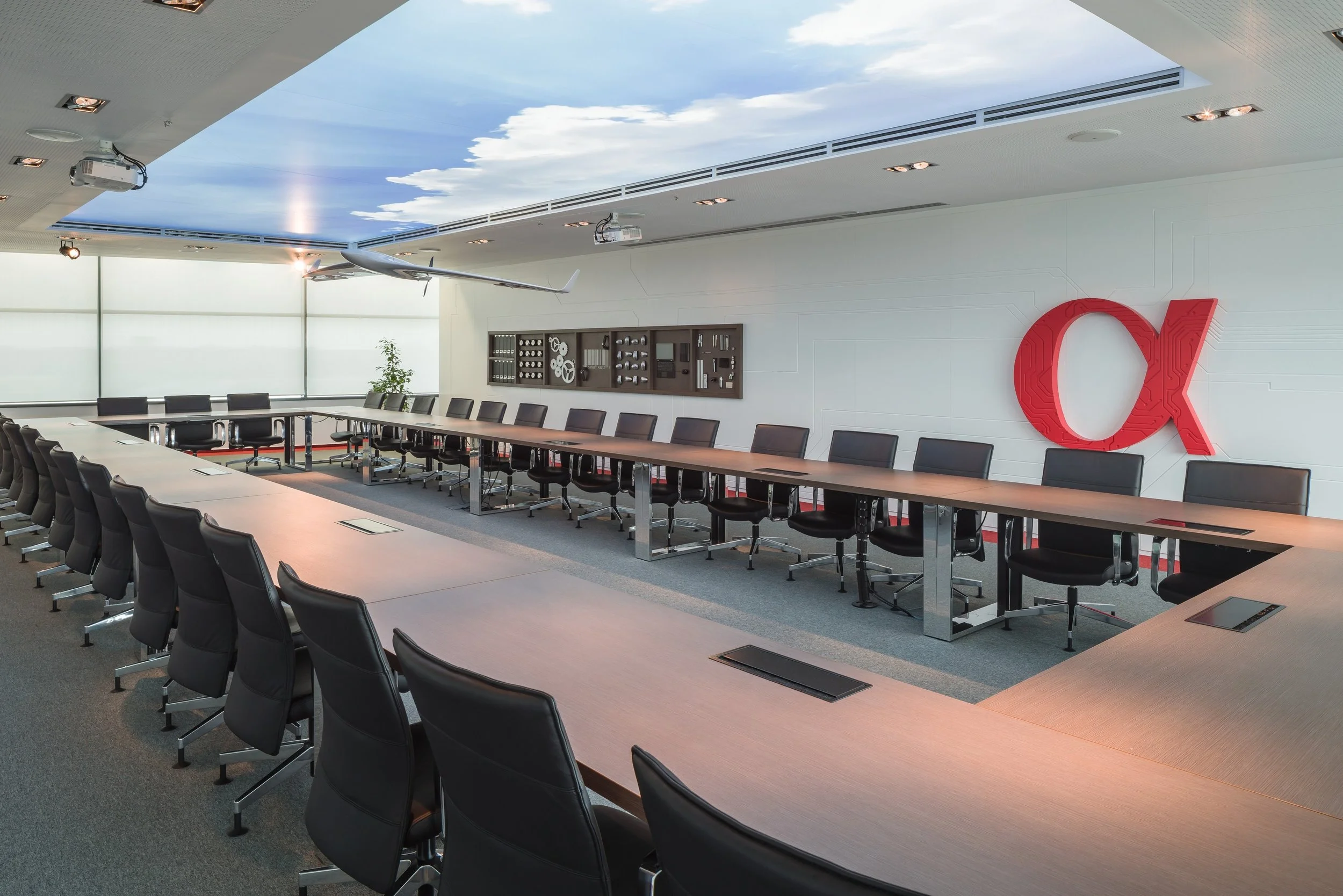 Modern conference room with U-shaped table setup, black office chairs, a ceiling mural of sky and clouds, large window, exhibit of drones on the wall, and a bright red 'OX' logo.