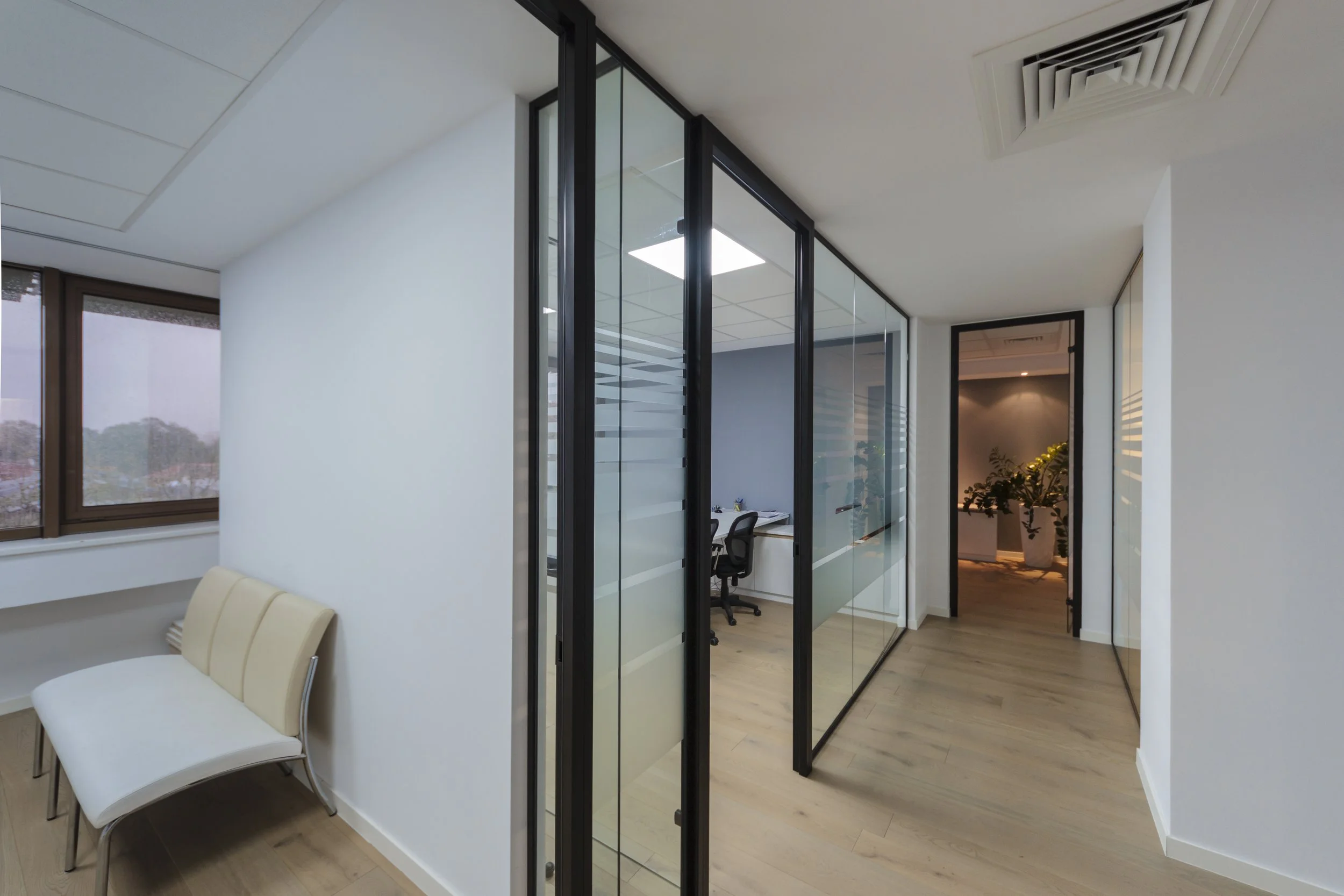 Modern office with glass-walled conference room, white walls, light wood flooring, and potted plants visible through a doorway.