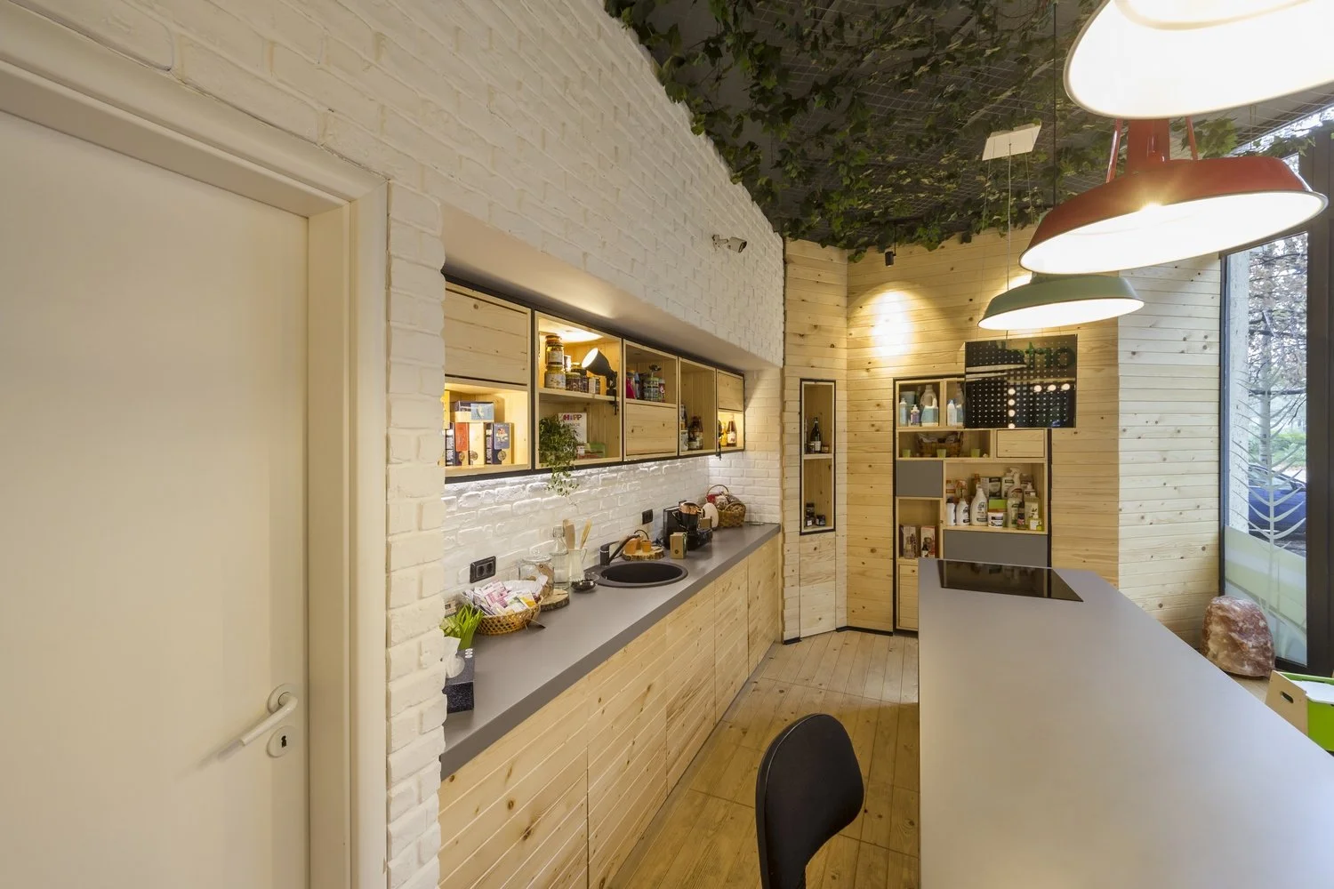Modern kitchen with light wood cabinets, white brick wall, and beige wood walls. Open shelving with food and kitchen supplies, hanging pendant lights, and large windows with a view of trees outside.