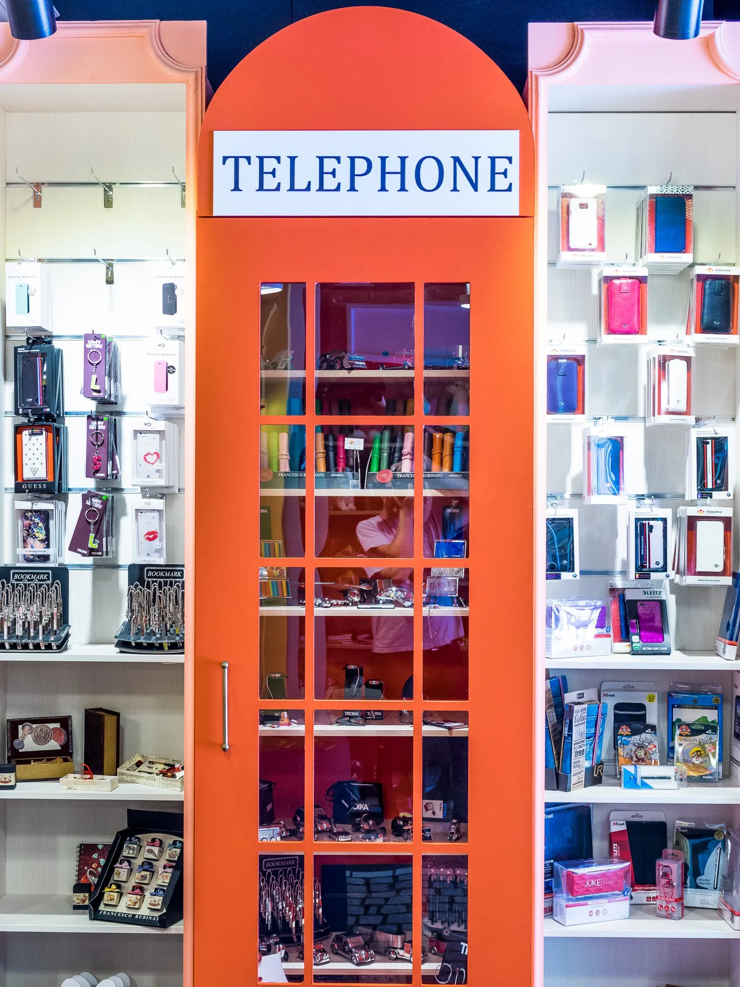 A display resembling a traditional red telephone booth filled with toy cars inside in a store, with shelves of phone cases and accessories on either side.
