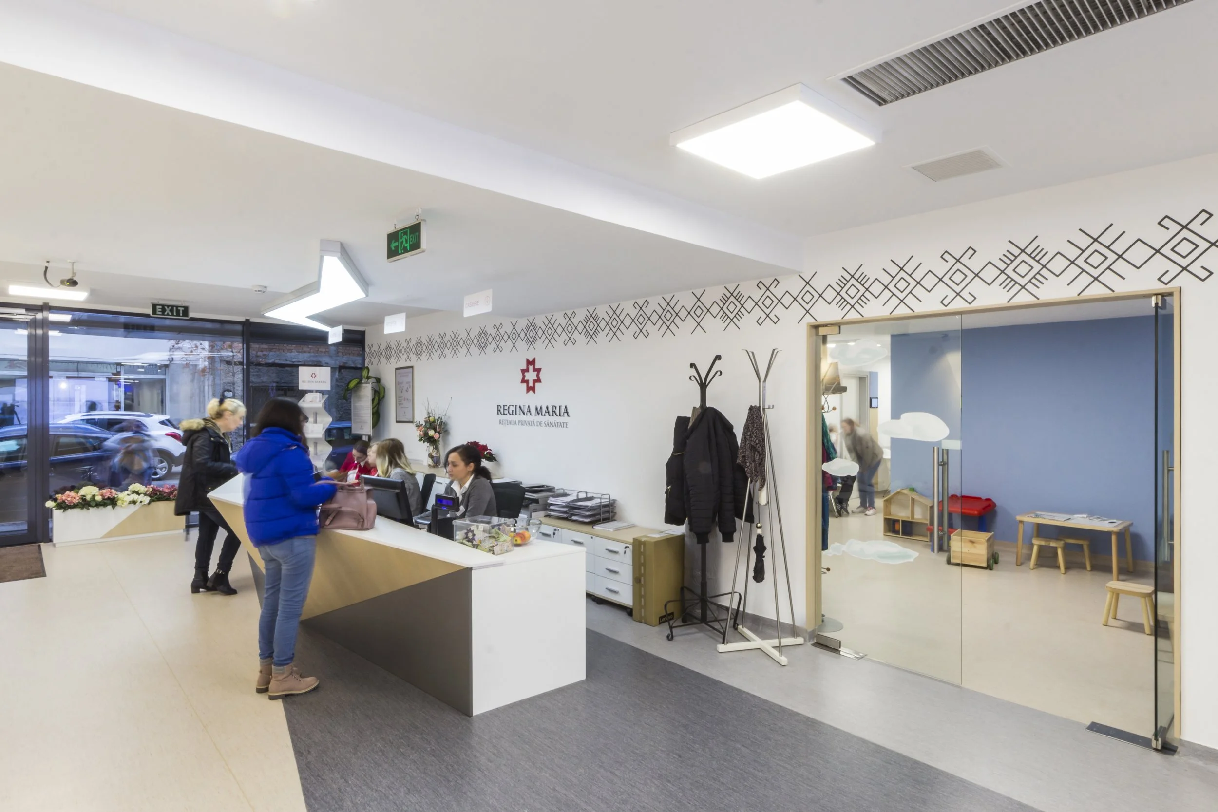 Reception area of Regina Maria clinic with a modern desk, staff members, visitors, coats on racks, and a playroom with toys visible through a sliding glass door.