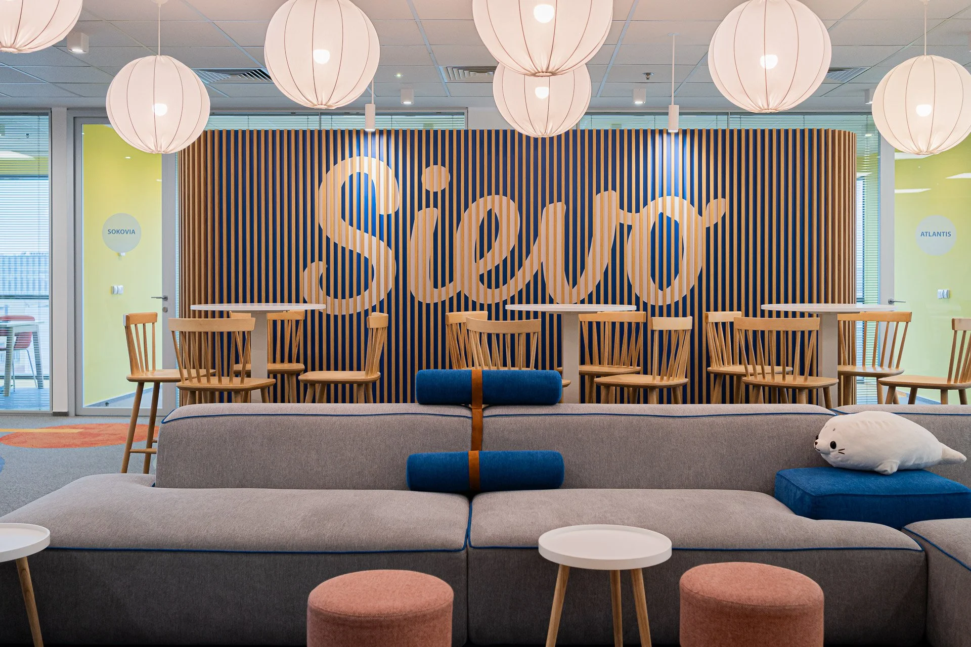 Modern lounge area with a large gray couch, blue headrests, and a lamp plush toy, in front of a bar with a wood slat backdrop that spells 'Sio' against a yellow wall with two doors labeled 'SOKOVIA' and 'ATLANTIS'.