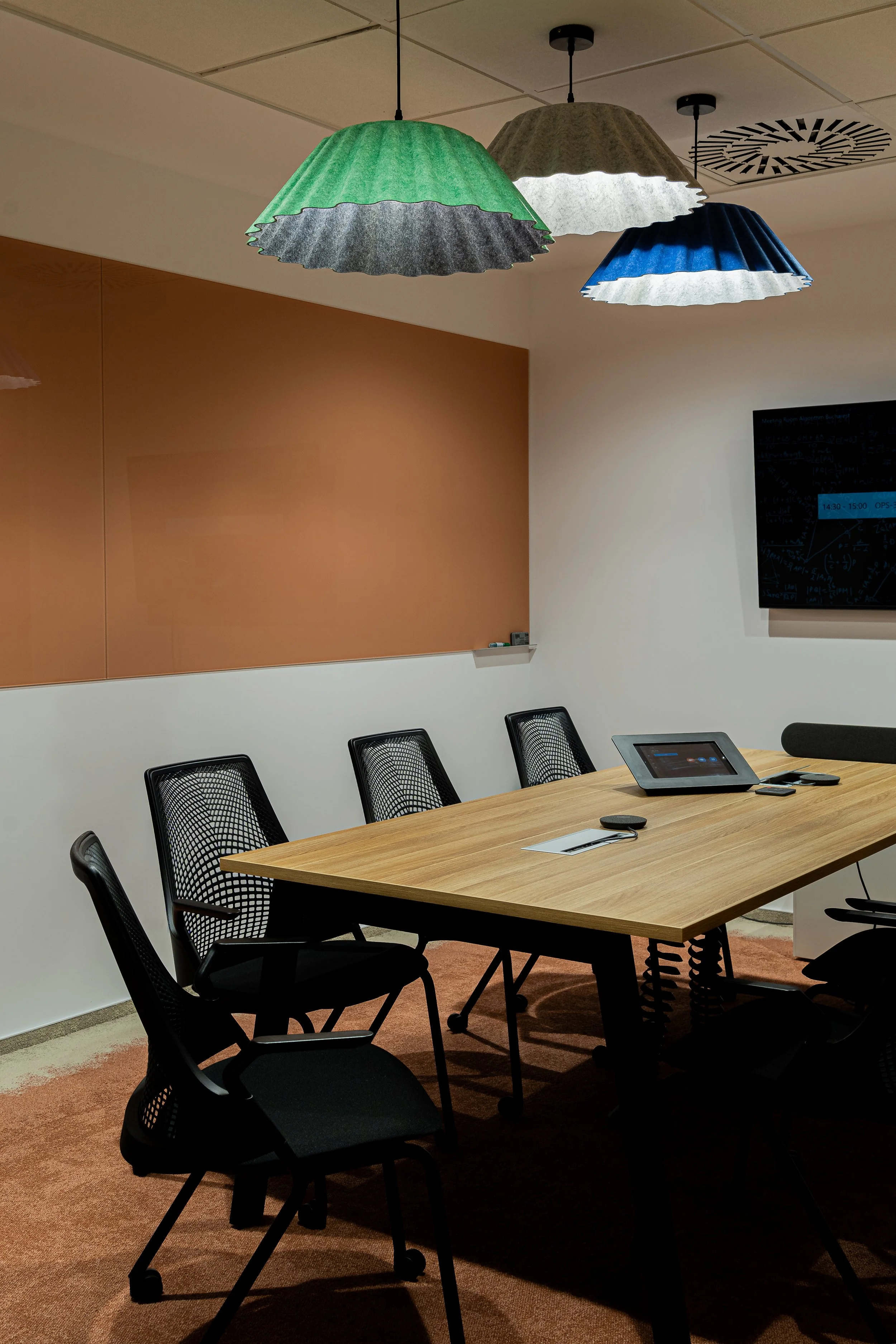 Meeting room with a wooden conference table surrounded by black chairs, three colorful fabric pendant lights hanging from the ceiling, a large screen on the wall, and a whiteboard on another wall.
