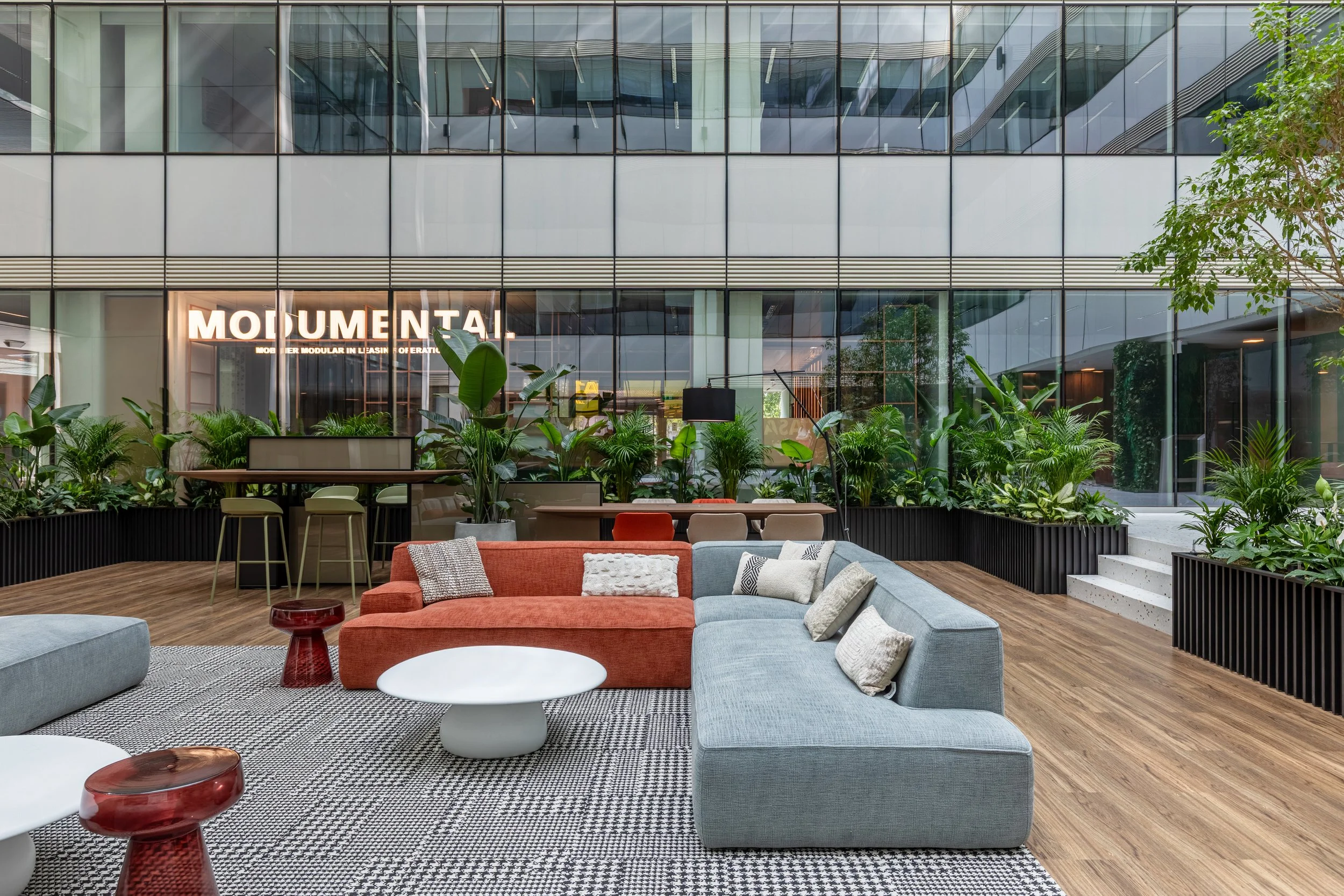 Modern office lounge with a sectional sofa in shades of grey, red, and beige, decorated with pillows, and round coffee tables. The lounge features a large window with lush green plants and a view of an office building with glass walls.