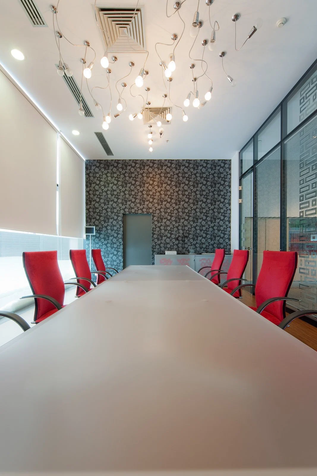 A modern conference room with a long white table, red chairs, decorative black and white wallpaper on the back wall, and a ceiling with multiple hanging light bulbs.