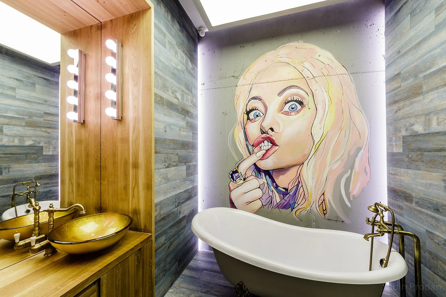 A bathroom with a large graffiti-style mural of a woman with blond hair, blue eyes, and pink lips on the wall behind a vintage clawfoot bathtub. The bathroom has wooden accents, including a wooden vanity with a golden sink, and wood-paneled walls.