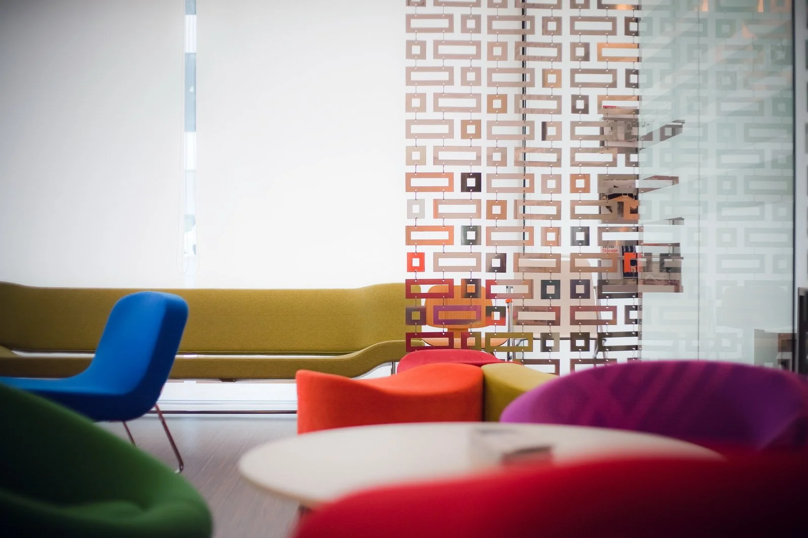 Colorful modern lounge area with various chairs and couches, a decorative room divider, and a window with vertical blinds.