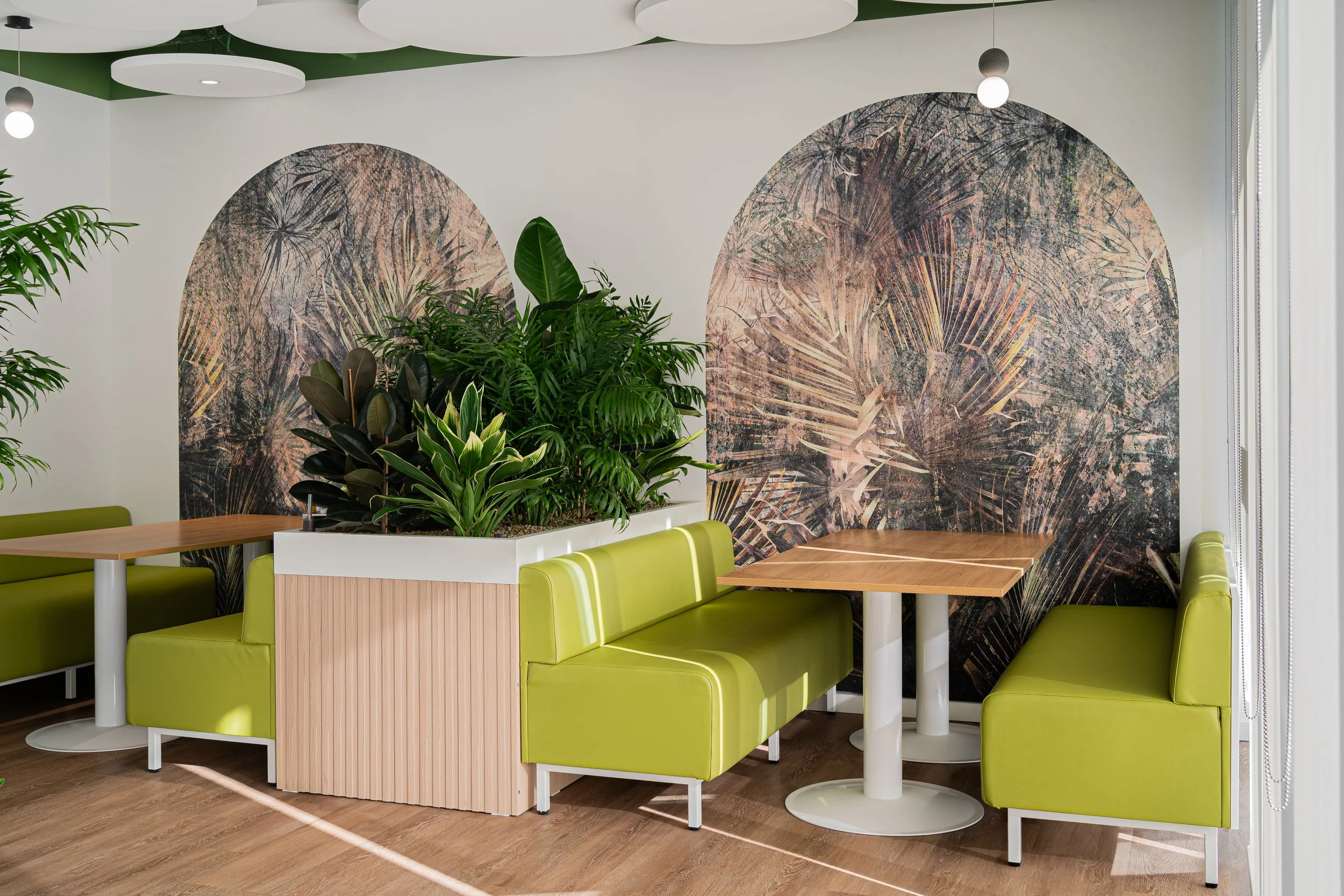 Interior of a modern cafe or restaurant with green cushioned booths, wooden tables, and large leafy green plants. The wall features a decorative mural with abstract botanical patterns, and there are hanging pendant lights.