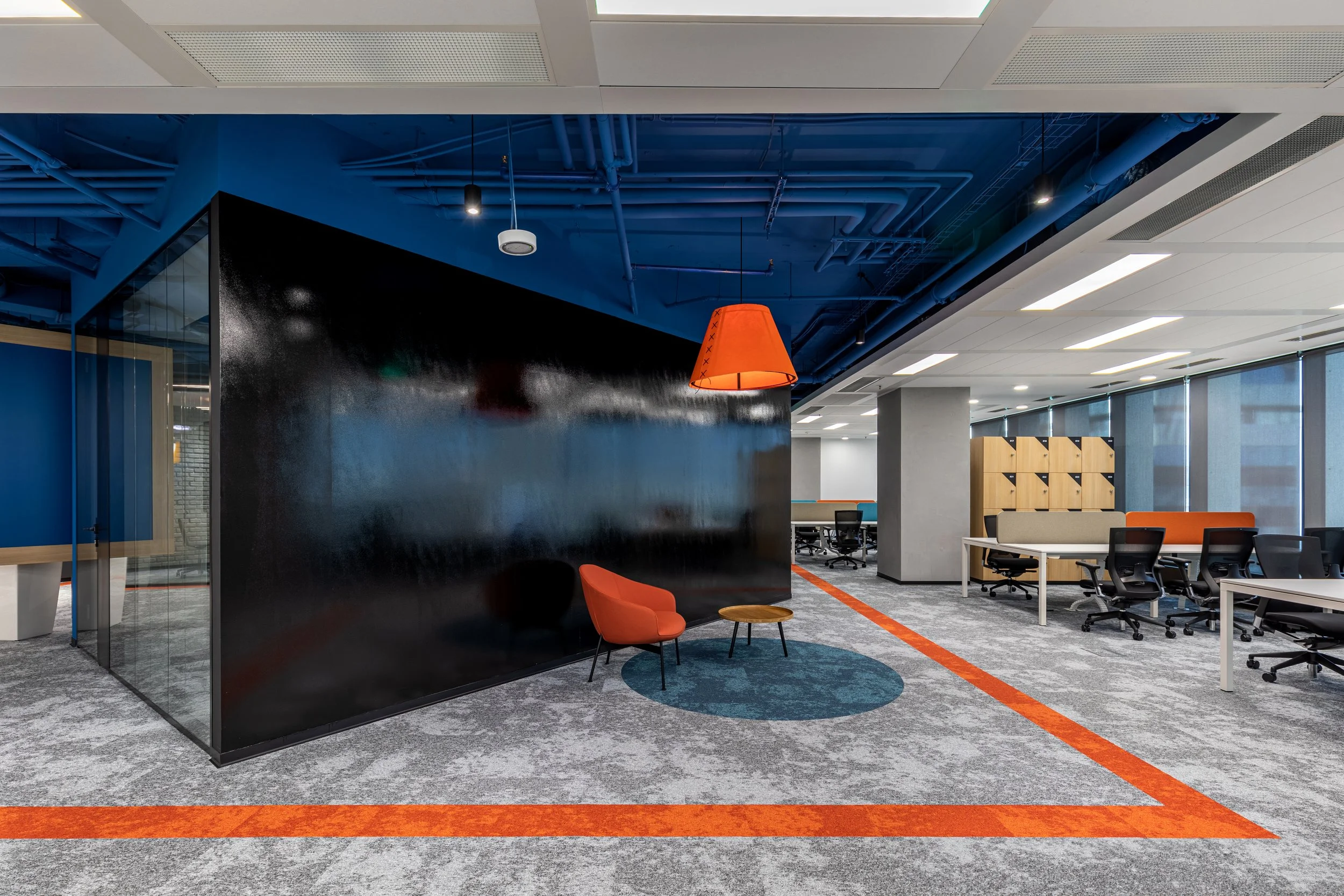 Modern office space with a seating area featuring an orange chair, orange lamp, and small round table, along with desks and chairs in the background under large windows.