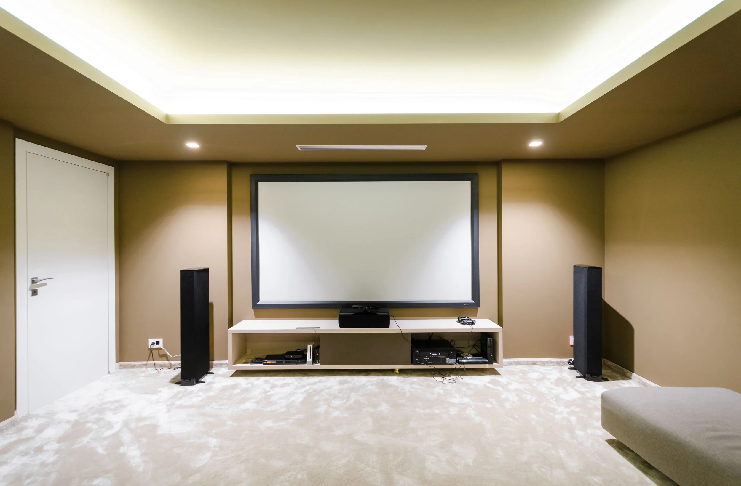 Home theater room with a large screen on a beige wall, two tall black speakers, a white entertainment unit, and a beige carpeted floor.