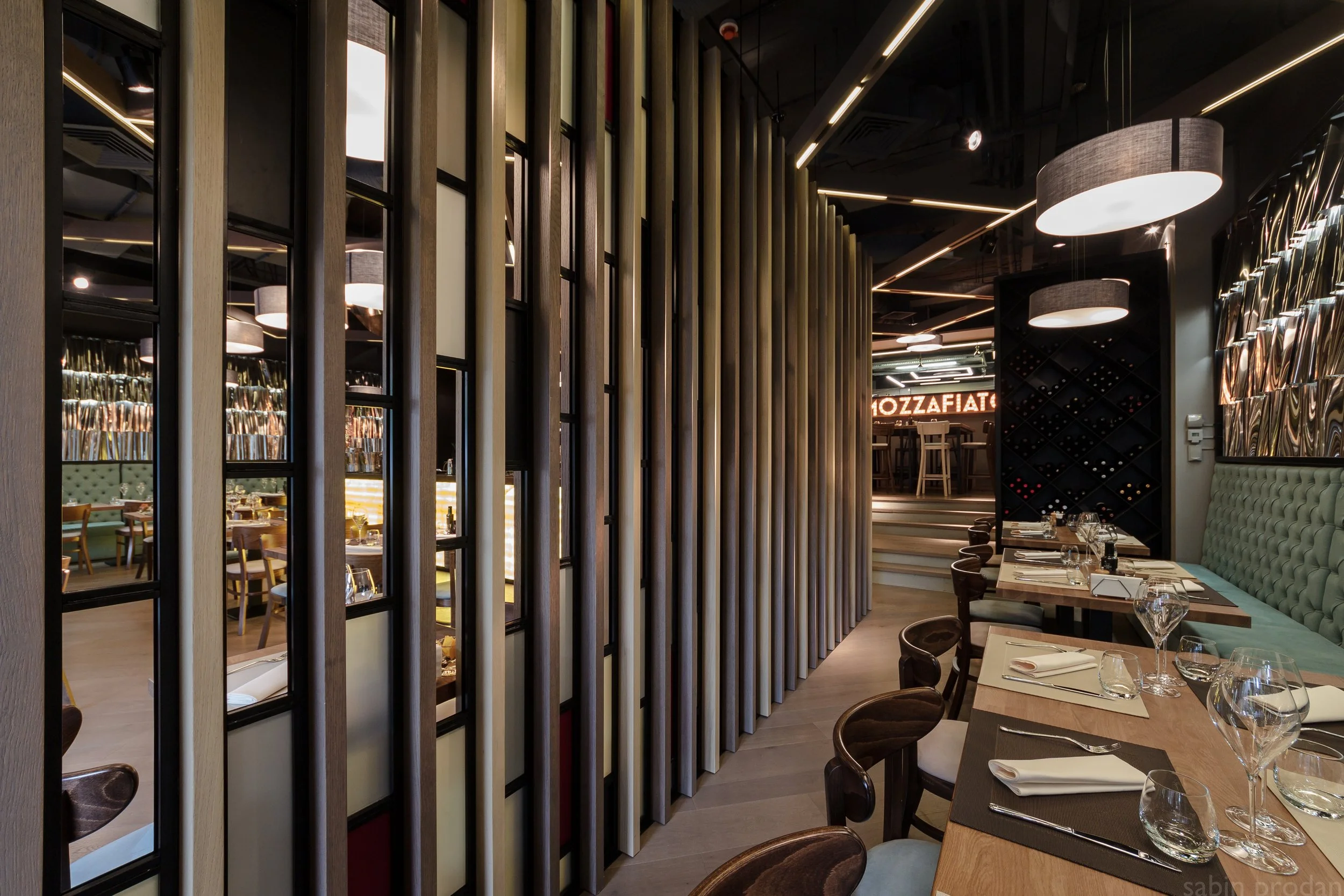 Interior of a modern restaurant with dining tables, chairs, decorative wall panels, and a wine rack in the background