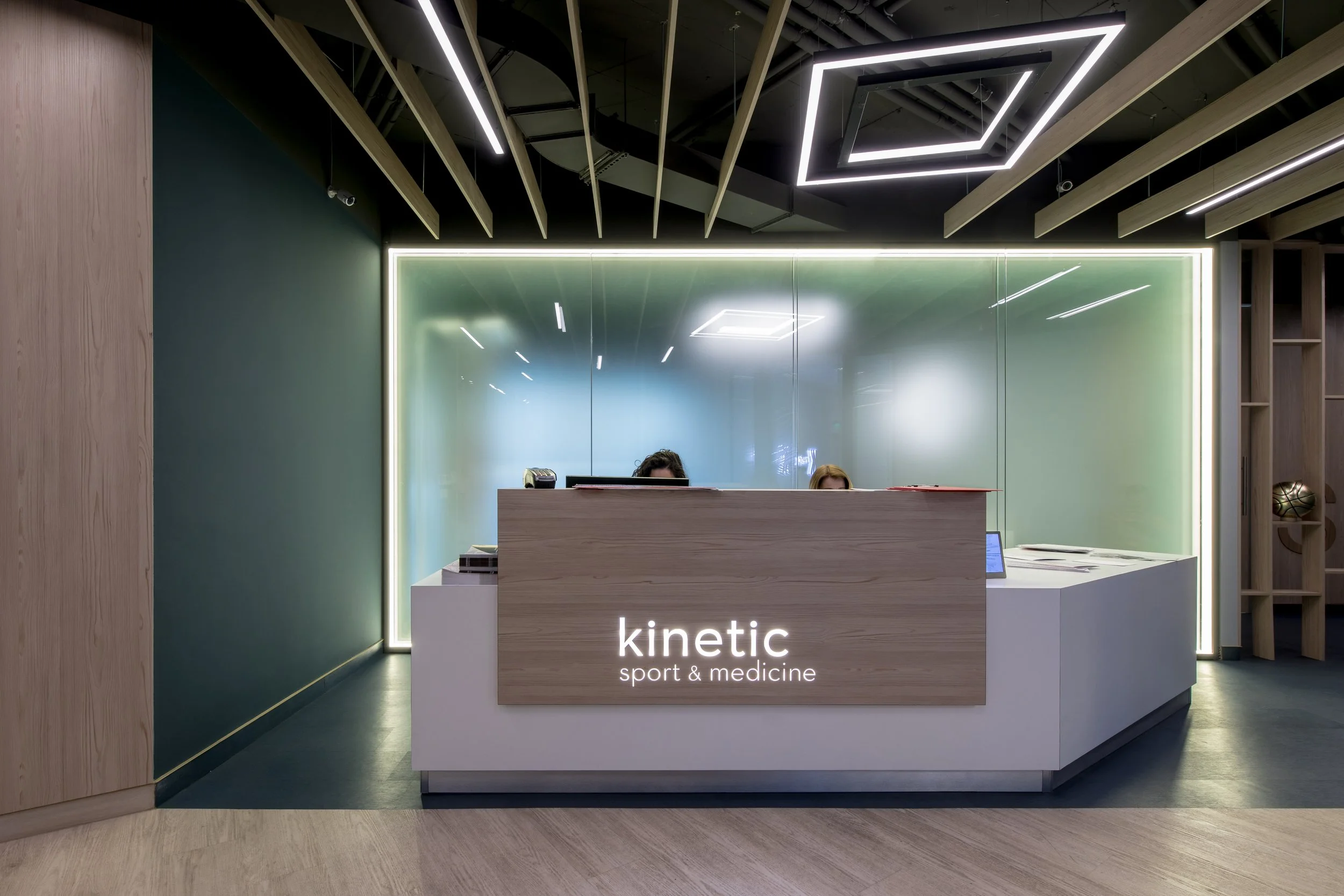 Modern reception desk with a wooden front panel, illuminated sign reading 'kinetic sport & medicine,' behind a glass wall with green tint, two receptionists partially visible, black ceiling with geometric LED lighting, wood and paneling decor.