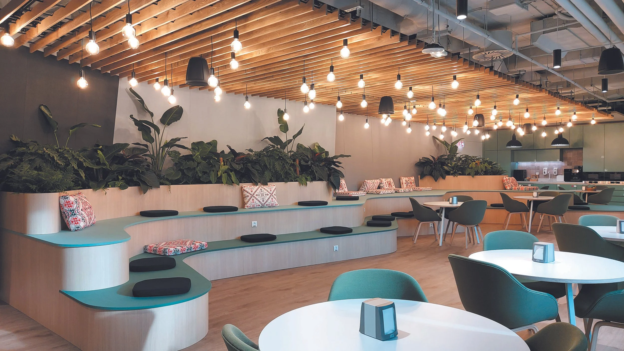 Modern restaurant interior with round tables, green and teal chairs, black cushions, and plants; wooden ceiling with hanging light bulbs.
