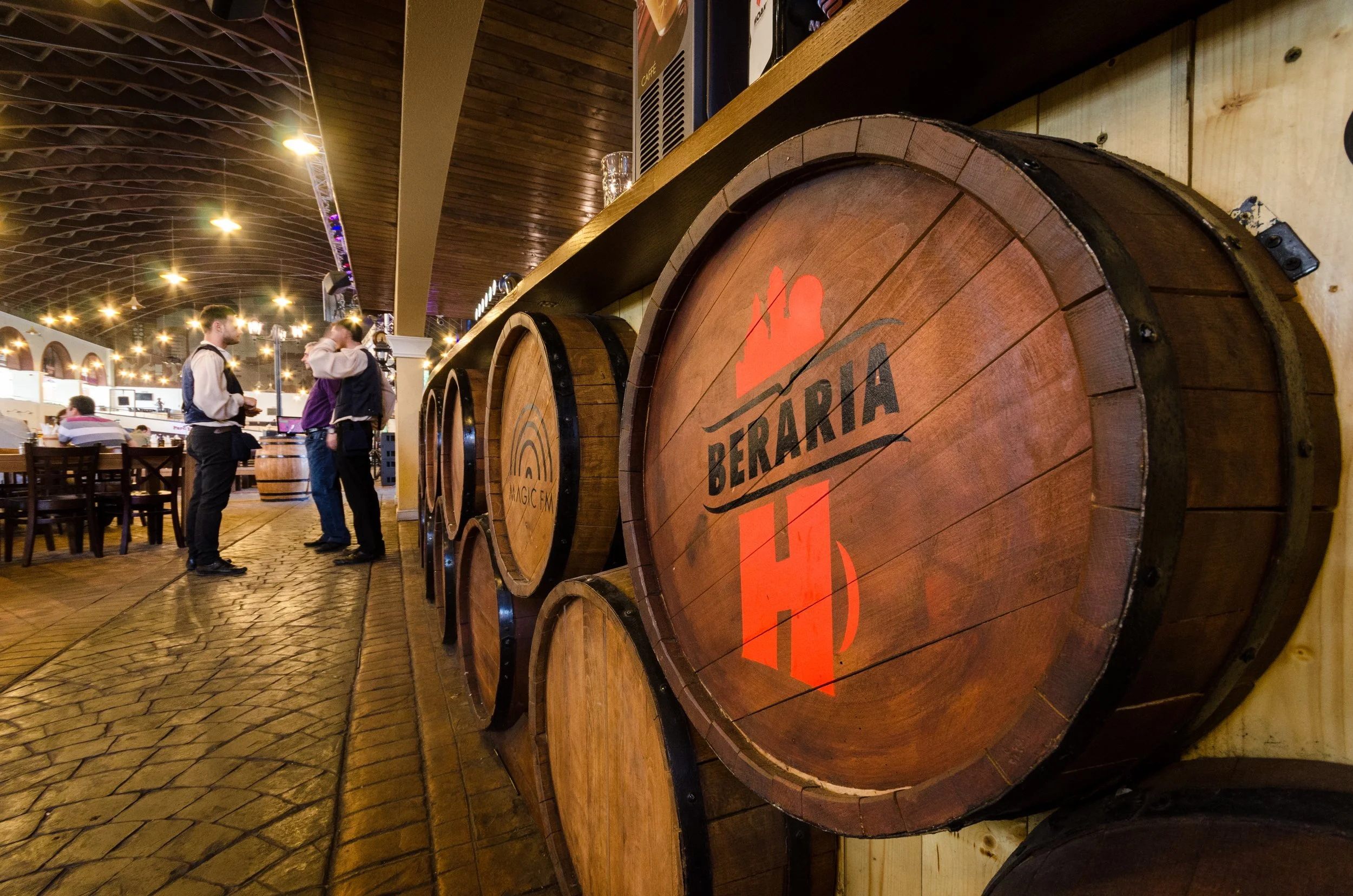 Large wooden barrels with brewery logos in a restaurant with several people standing and talking in the background.
