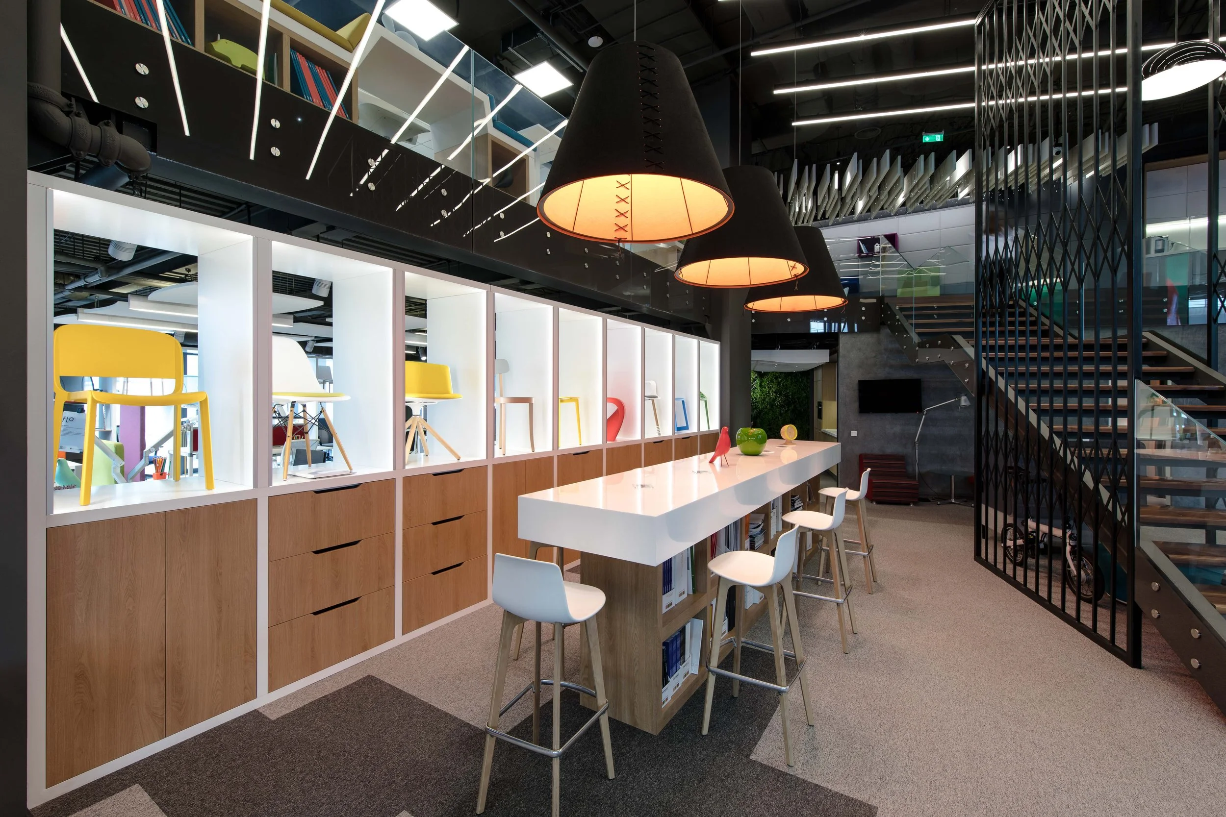 Modern office space with a white high-top table, white and wooden stools, colorful decorative objects, illuminated display shelves with chairs, black pendant lights, stairs with wooden steps, and a glass wall with a staircase and seating area in the background.