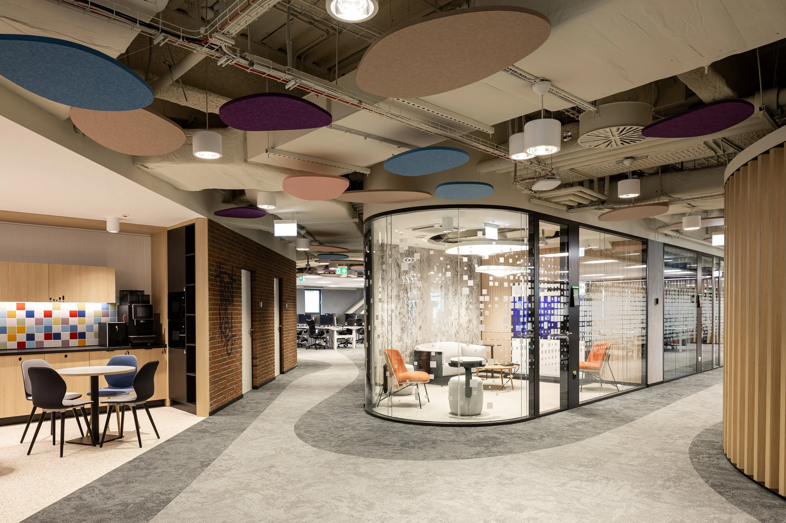 Modern office space with curved glass walls, orange and black chairs, a round table, and colorful ceiling panels in blue, pink, and purple. Open workspace with visible computer desks in the background.
