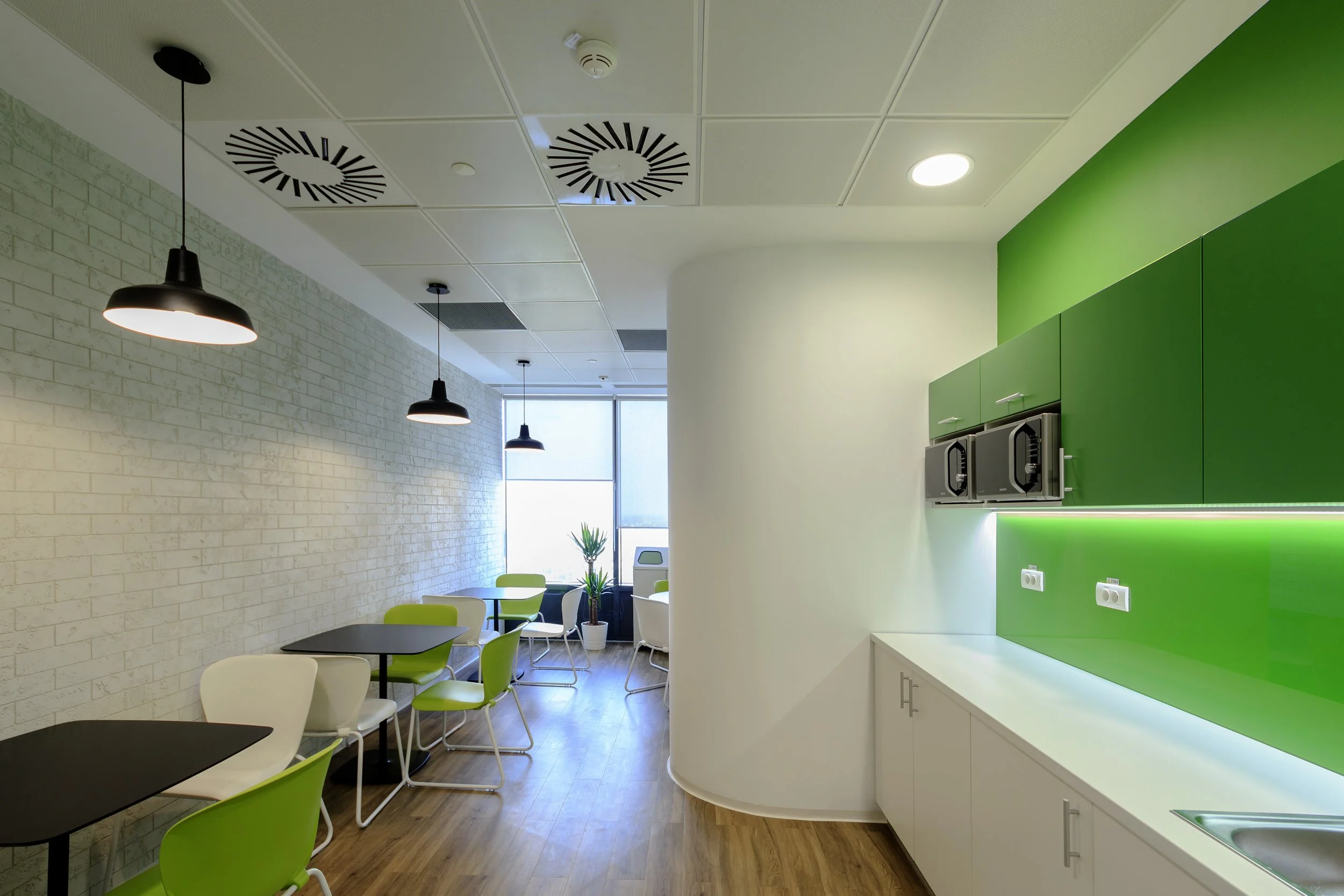 Interior of a modern workspace or break room with white brick wall, black tables, white and green chairs, pendant lighting, green cabinets, and a white counter with a sink.