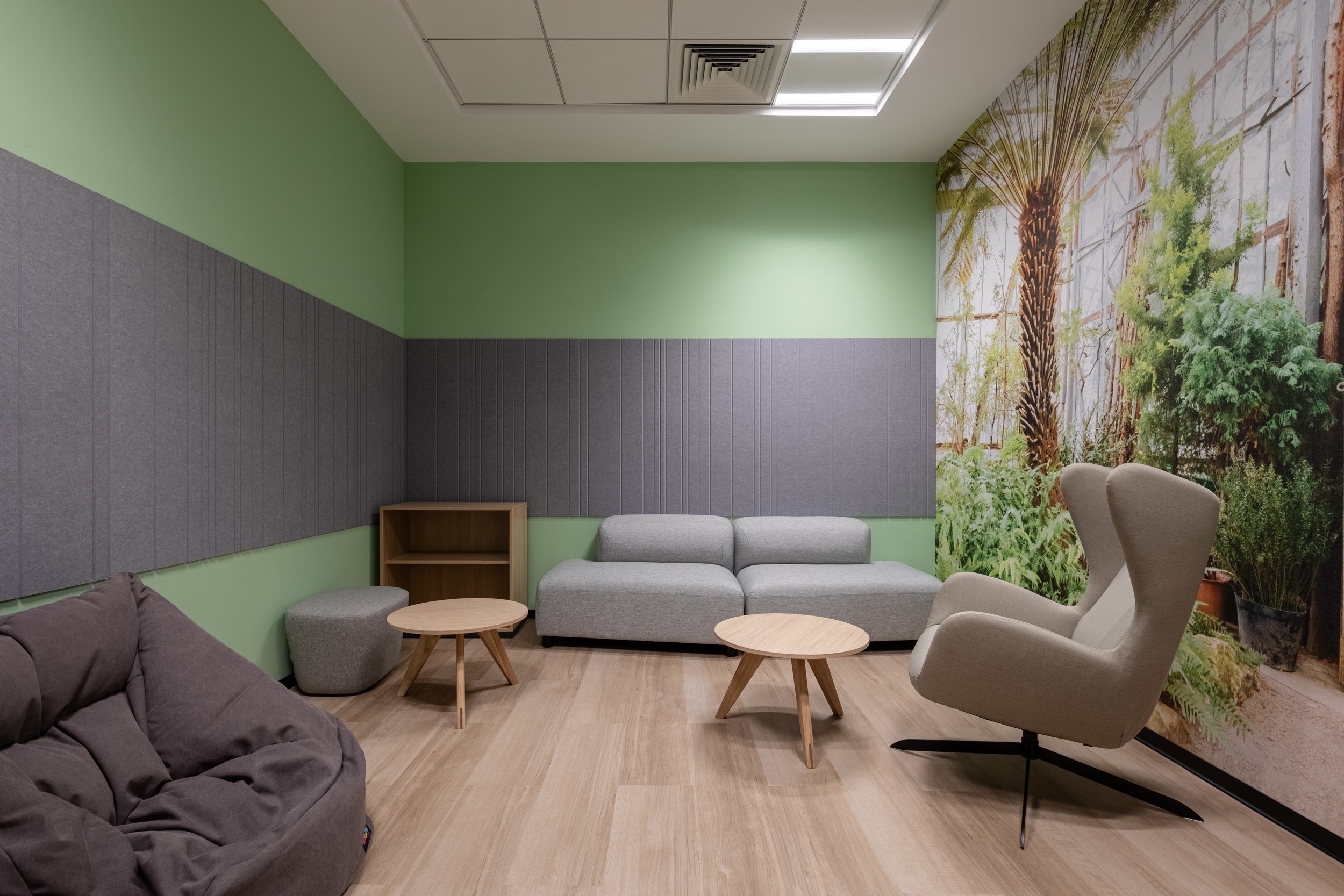 A cozy waiting room with green and gray walls, a gray couch, a white armchair, small wooden coffee tables, a wooden shelf, and a large nature-themed mural.