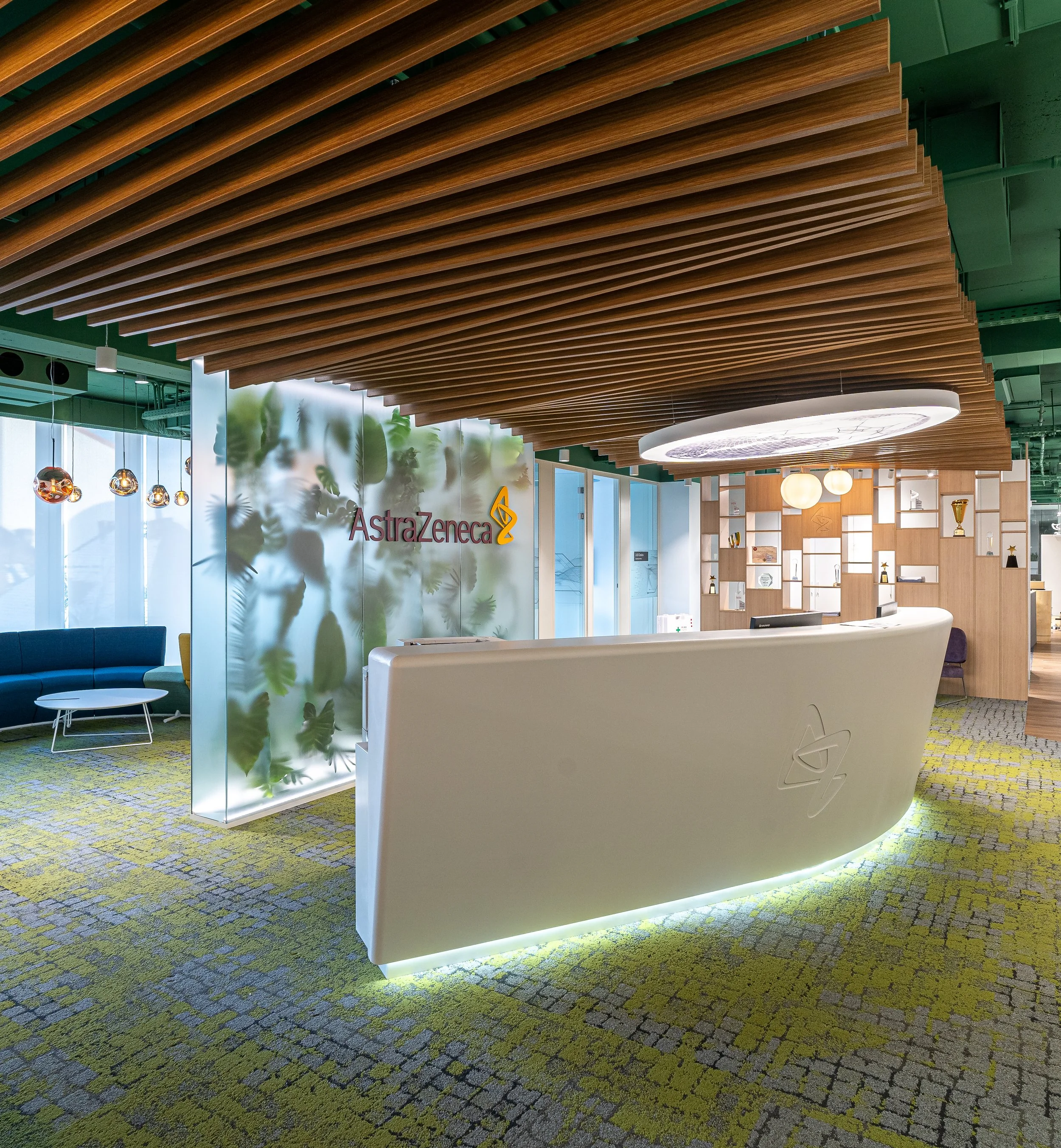 Modern office reception area with a white curved desk, wooden ceiling design, and green patterned carpet. The walls feature a glass panel with the AstraZeneca logo, and there are seating areas with colorful chairs and tables.