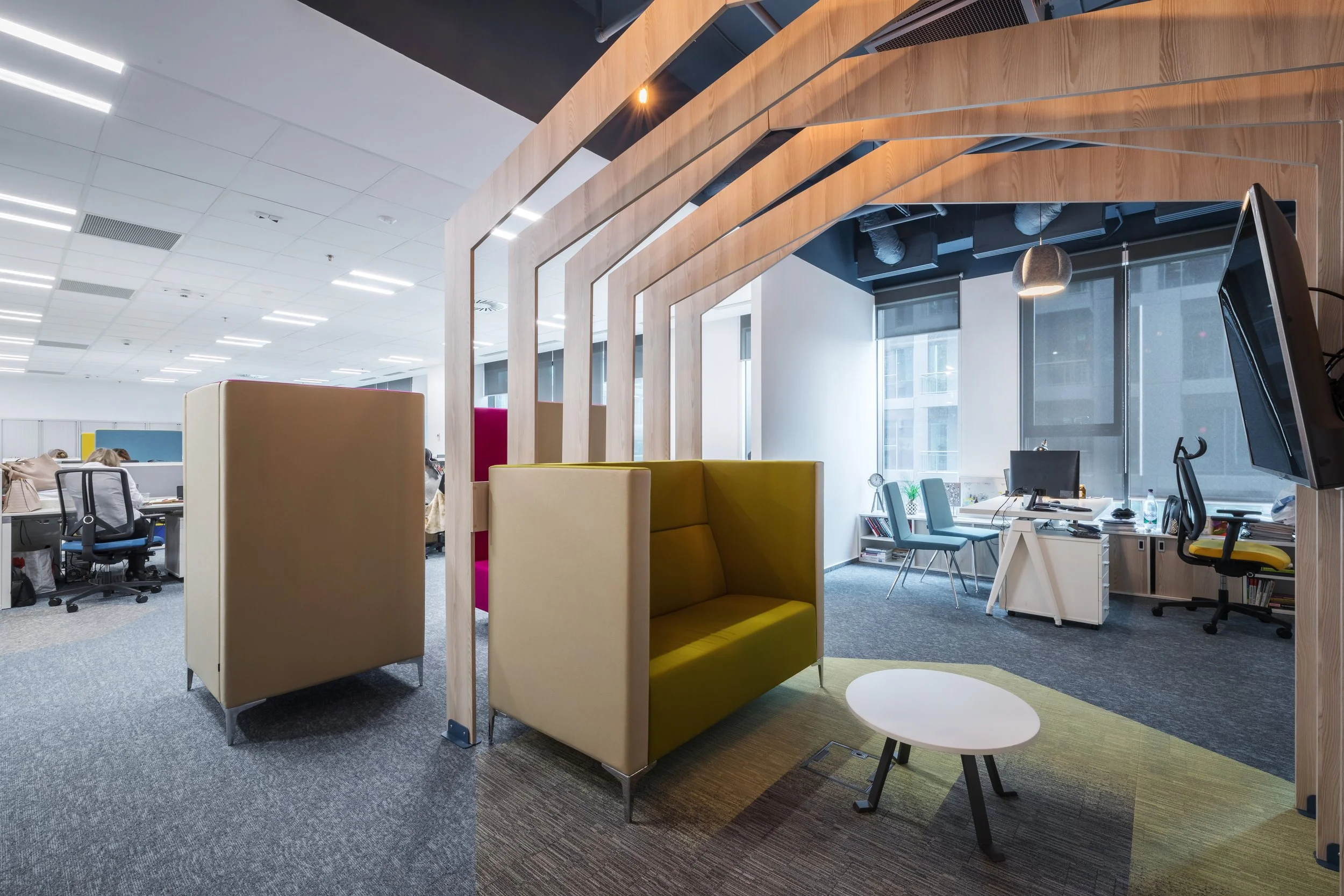 Modern office space with workstations, chairs, and a sitting area with colorful high-backed booths, a small white coffee table, and open work areas near large windows.