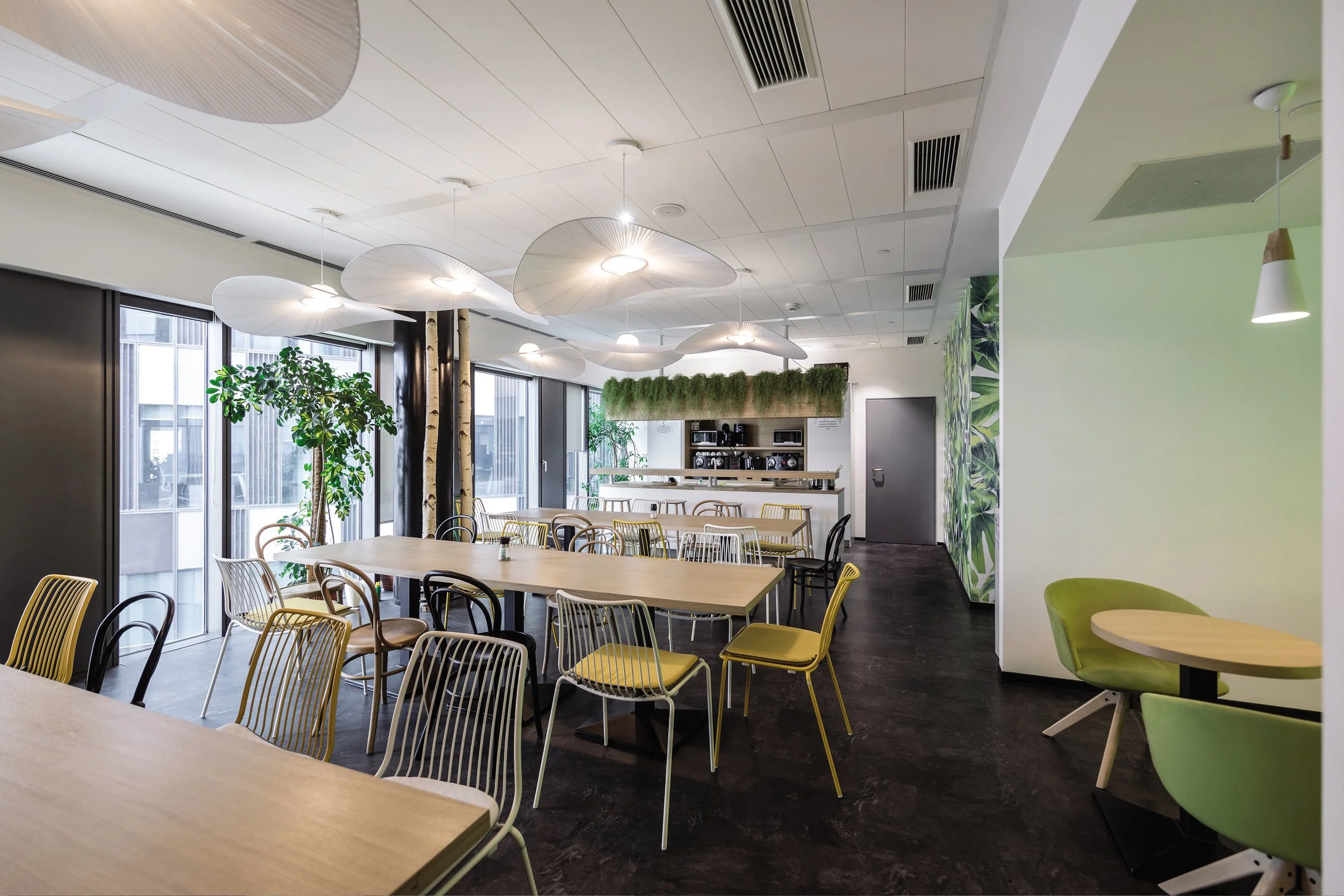 Modern cafe or lounge with light-colored tables, mixed chairs, large windows, indoor plants, and contemporary ceiling lights.