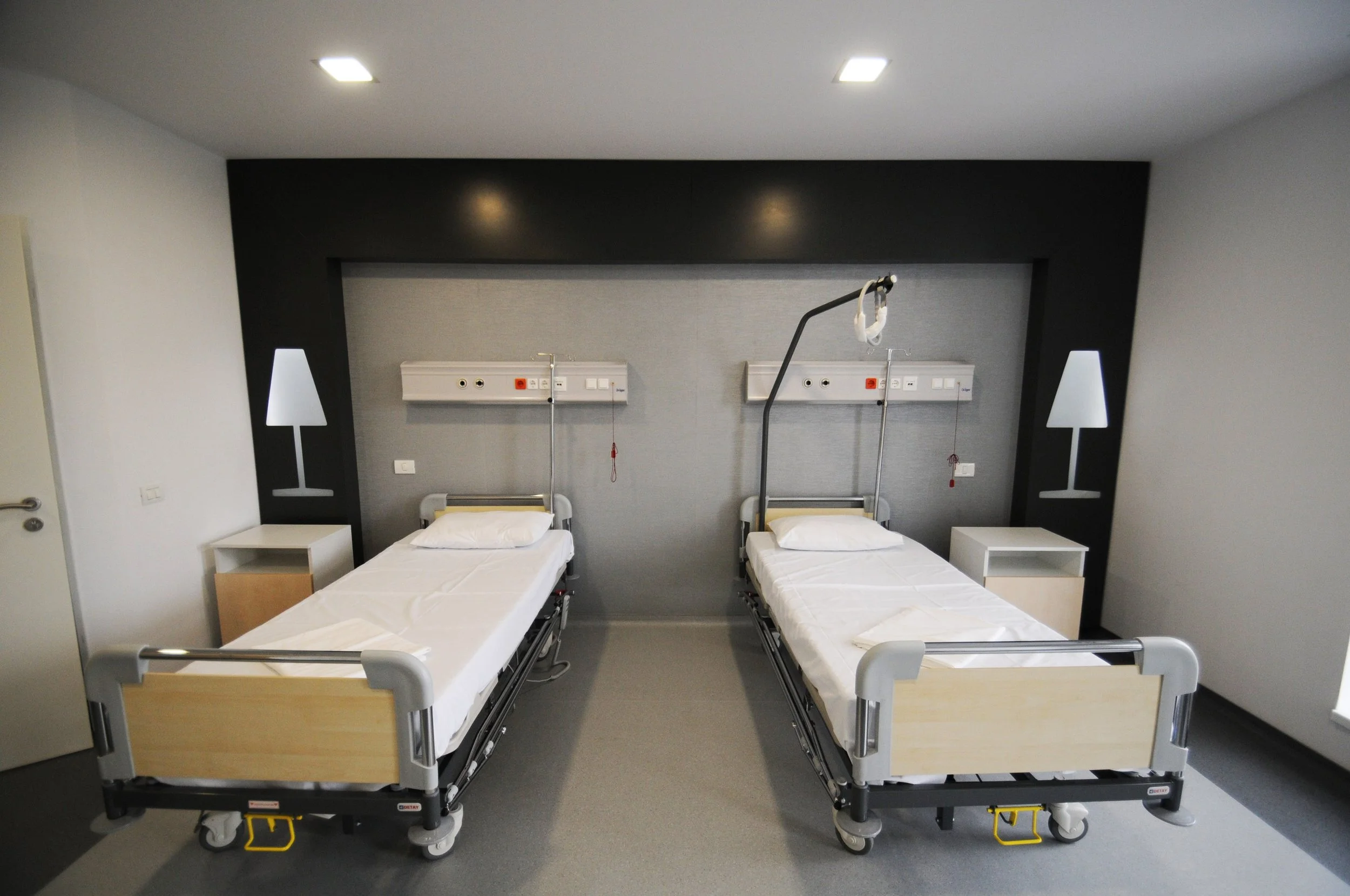 Two hospital beds with white sheets and pillows, placed side by side in a patient room. Each bed has a bedside table and attached medical equipment and outlets on the wall behind. The room has a black accent wall with lamps on either side.