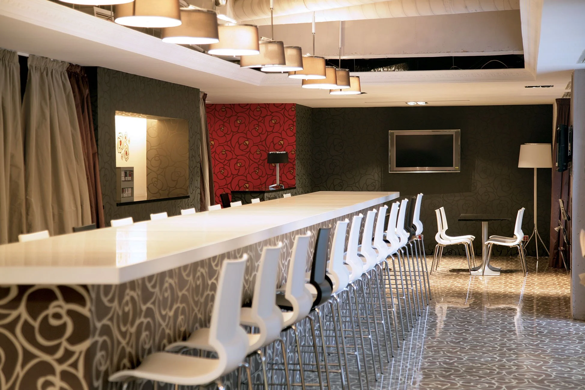 Modern hotel lounge with a long white bar counter, white bar stools, a wall-mounted TV, a small round table with white chairs, and stylish lighting fixtures.