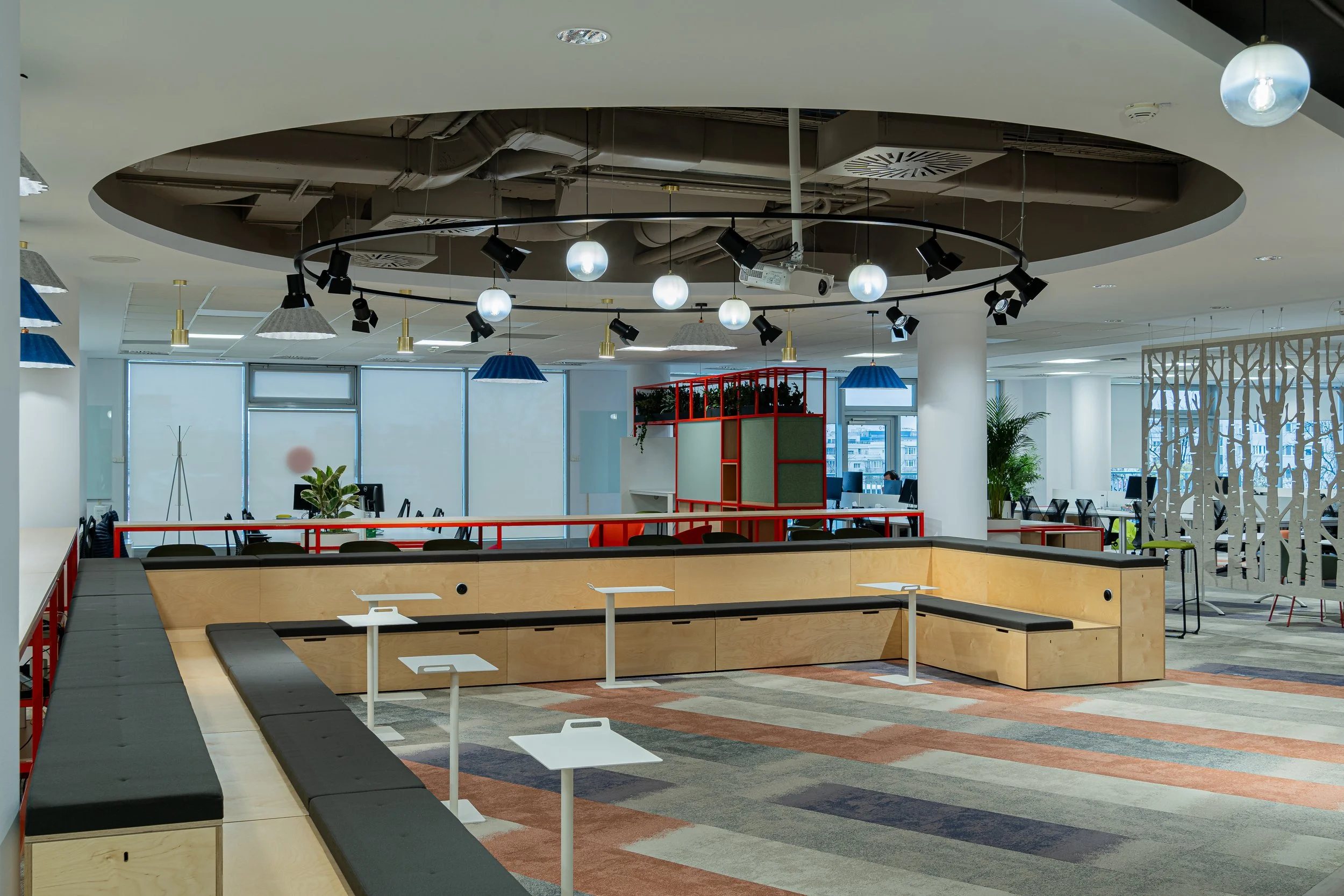 Modern open-concept office space with seating areas, desks, plants, and ceiling lighting fixtures.