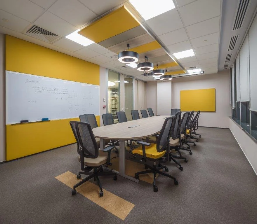 A modern conference room with a long oval table and 10 office chairs, black with yellow seats, arranged around the table. The room has a whiteboard on the left wall, a yellow panel above it, and a yellow rectangular corkboard on the opposite wall. The ceiling features circular, modern light fixtures, and the room has large windows on the right side with blinds.