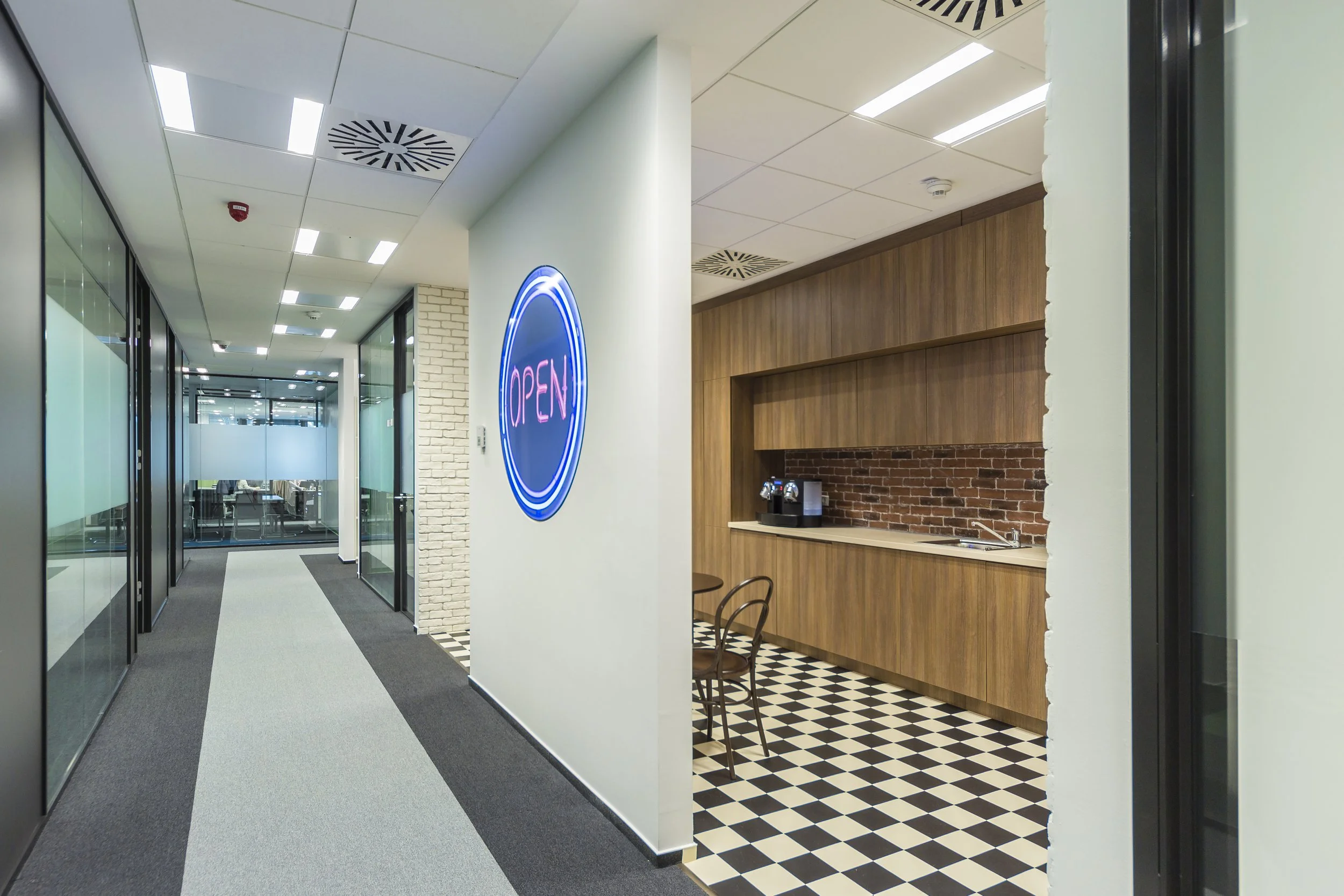 Modern office hallway with glass-walled conference rooms on the left, white walls, and a carpeted floor. An illuminated neon sign displaying 'OPEN' is mounted on the right wall near a small kitchenette with a checkered black-and-white tile floor, wooden cabinetry, a coffee machine, and two chairs.