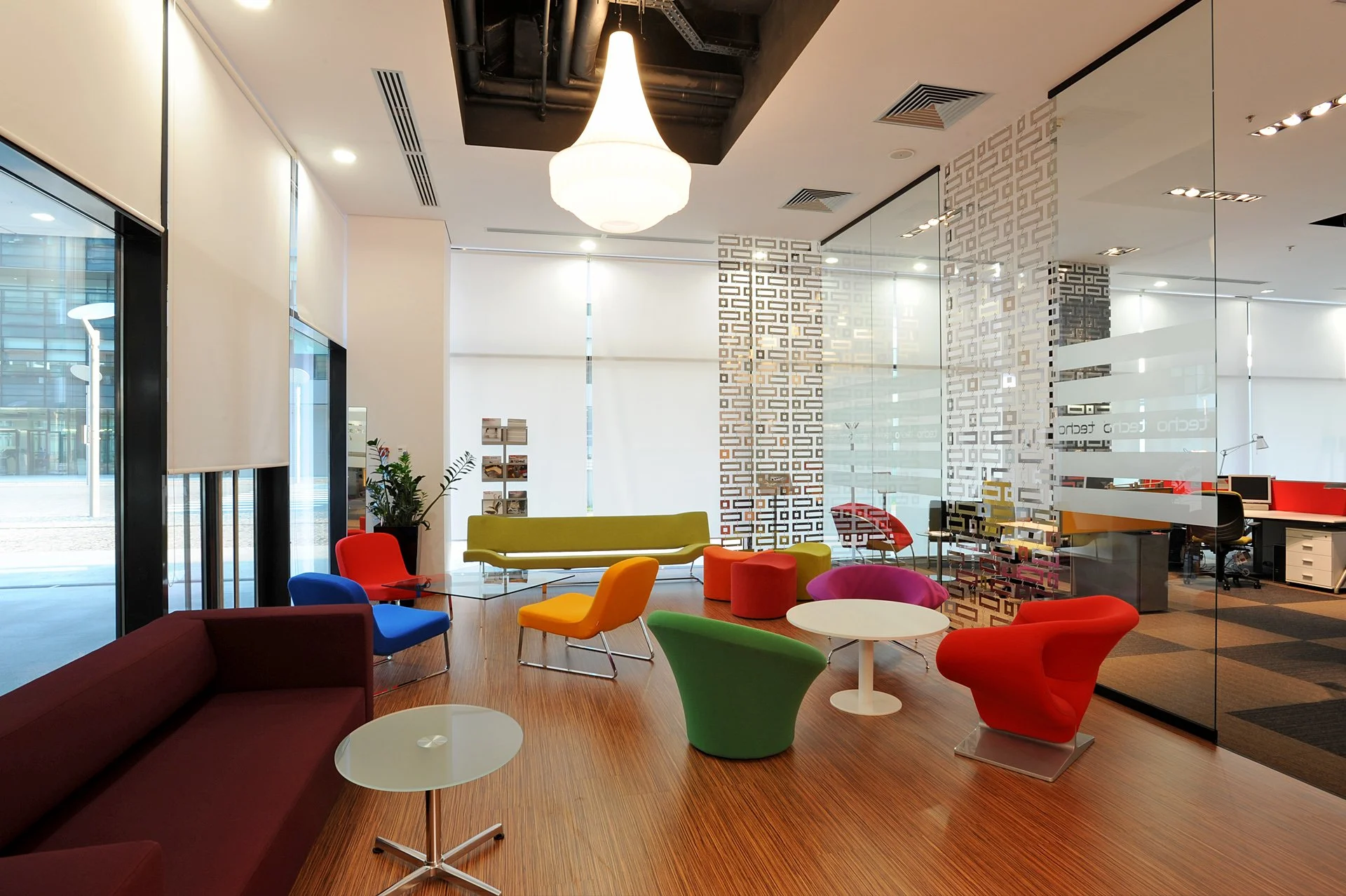 Colorful modern office lounge area with various chairs, a sofa, small tables, and a decorative partition, with glass walls and a chandelier overhead.