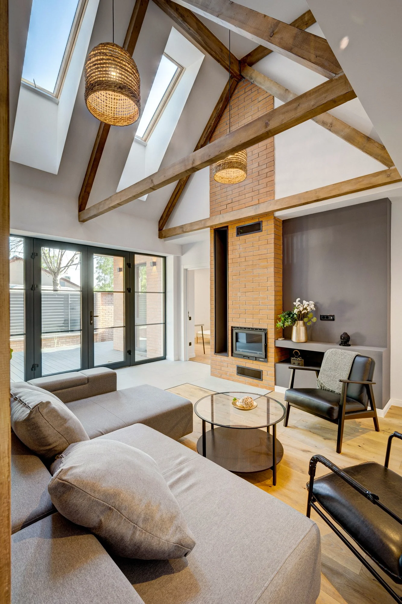 Modern living room with high vaulted ceiling, exposed wooden beams, skylights, a brick chimney, large sliding glass doors, a beige sectional sofa, a black armchair, round glass coffee table, and decorative items.