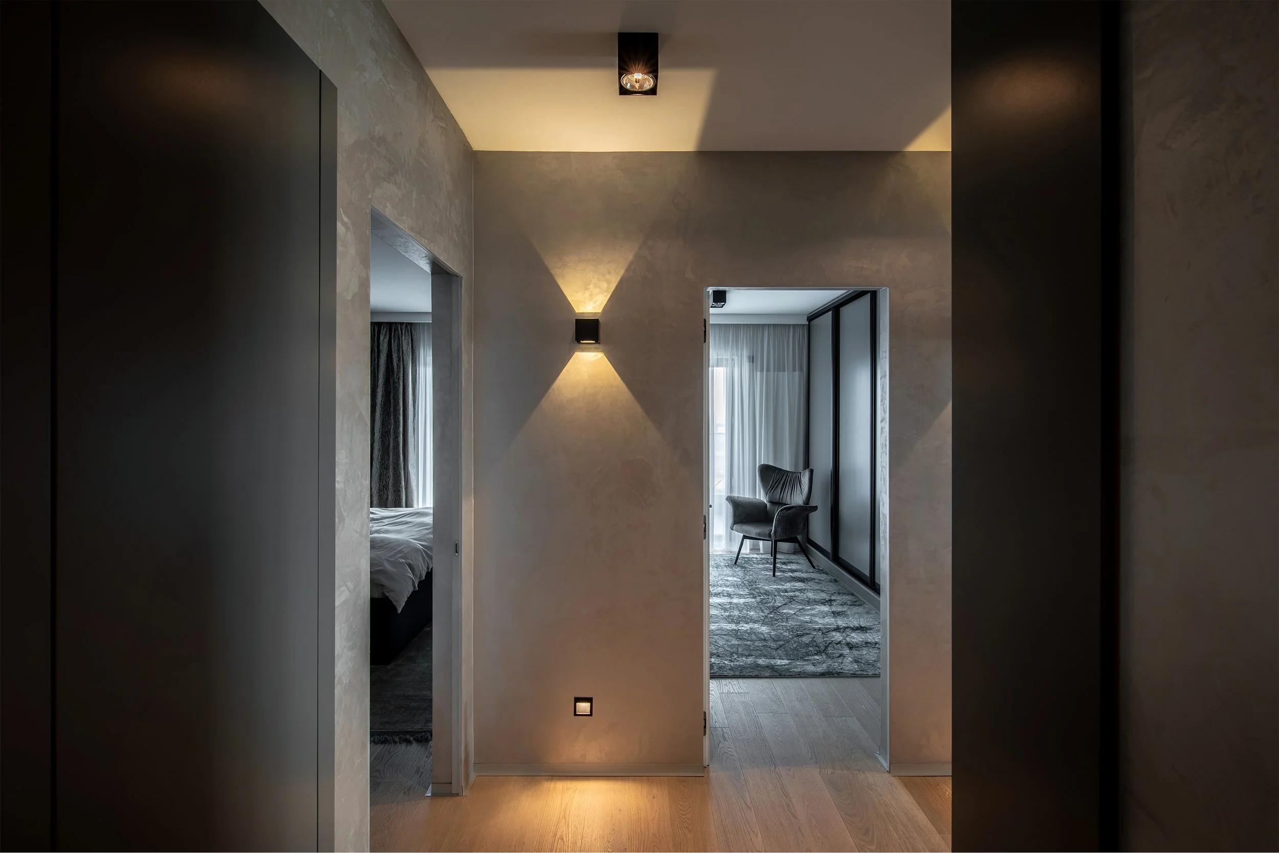 A modern hallway with beige textured walls, wooden flooring, and wall-mounted black light fixtures. The hallway opens to a bedroom on the left and a sitting area with a black armchair and a large window on the right.