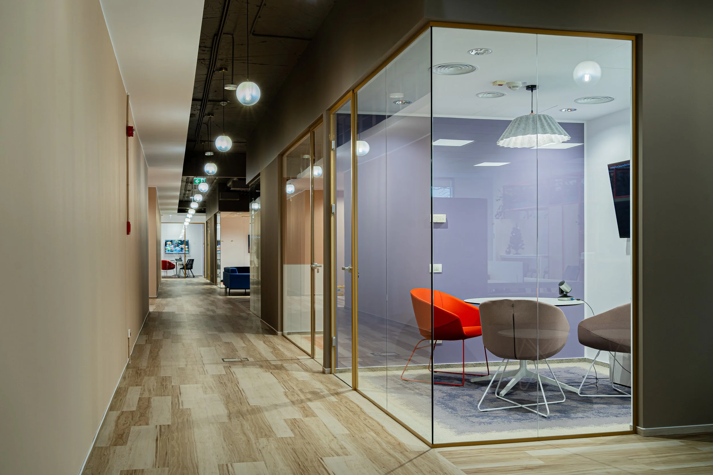 Modern office hallway with glass-walled meeting rooms on the right, illuminated by hanging pendant lights, with a view of a colorful meeting room interior featuring chairs and tables.