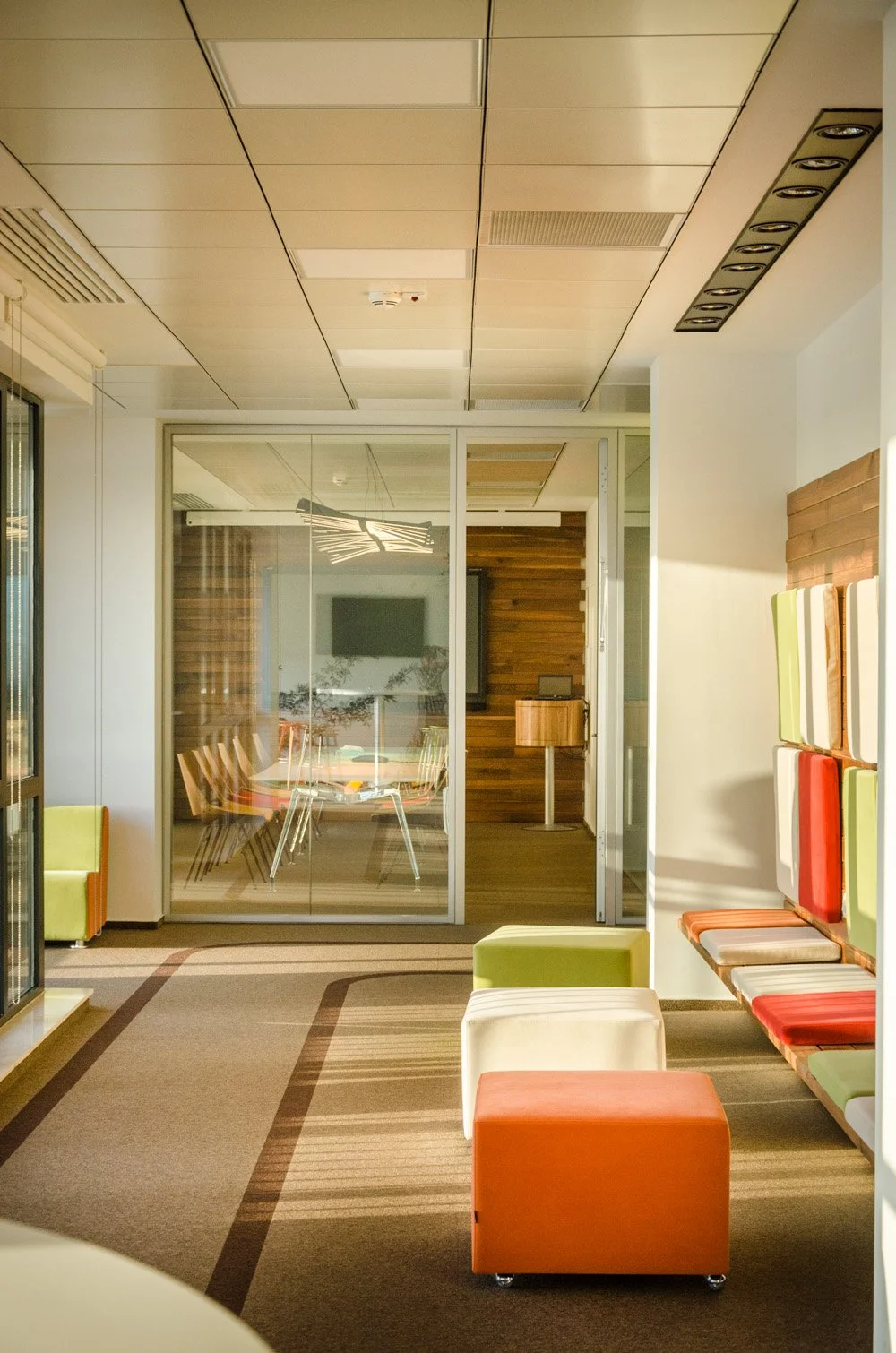 Modern office lounge area with colorful seating, a glass-walled conference room, and wood-paneled walls.