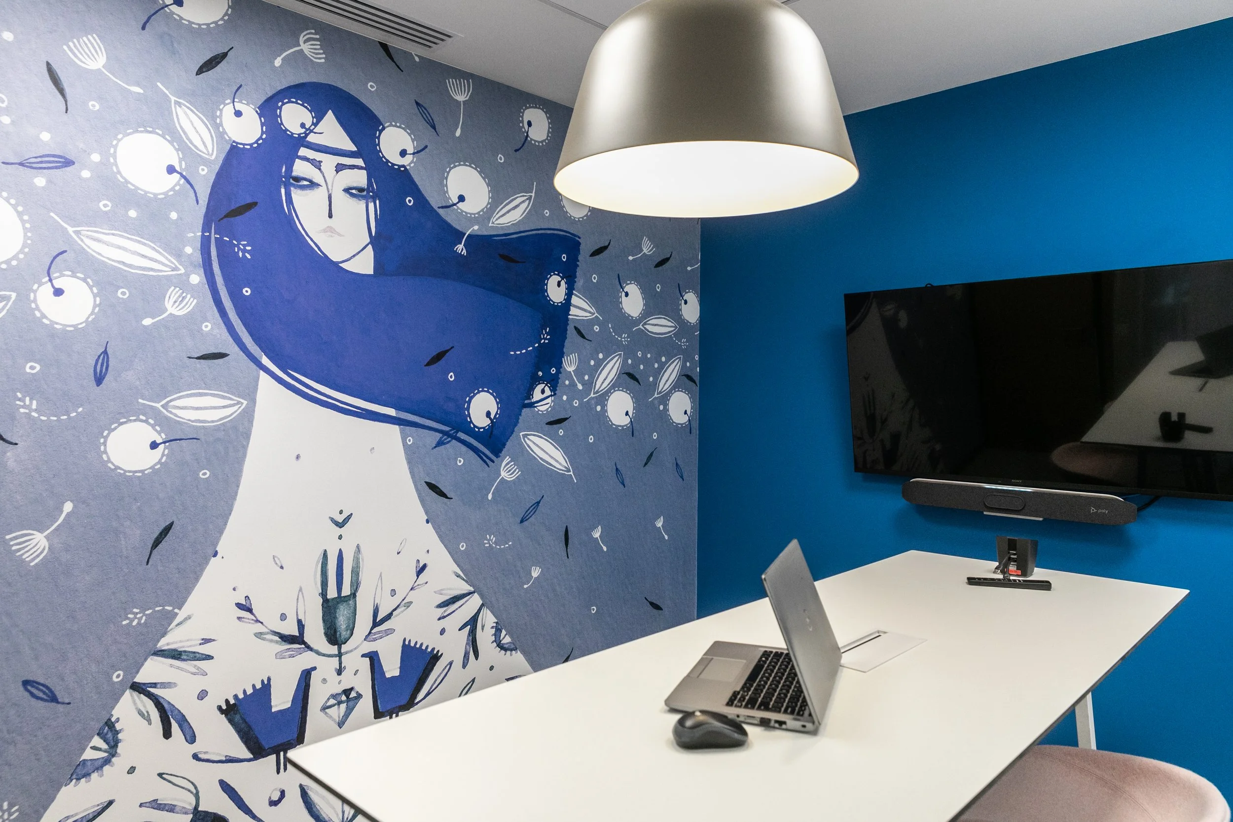 Office room with a large wall mural of a stylized woman in blue tones, a white conference table with a laptop and mouse, a wall-mounted TV, a dark grey speaker, and a white pendant light.