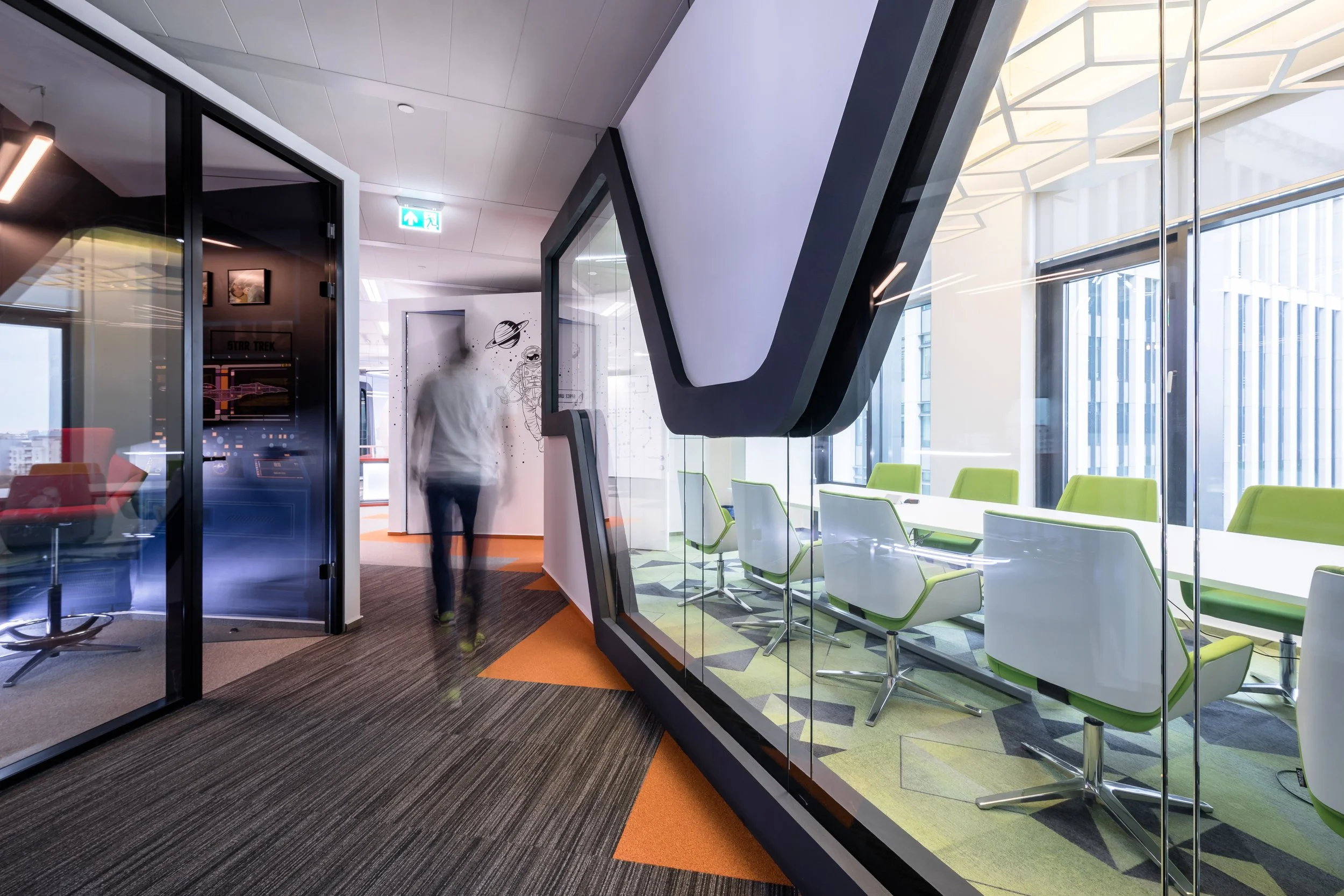Modern office space with glass-walled meeting rooms, green and white chairs around a conference table, and futuristic wall art of planets and space exploration.