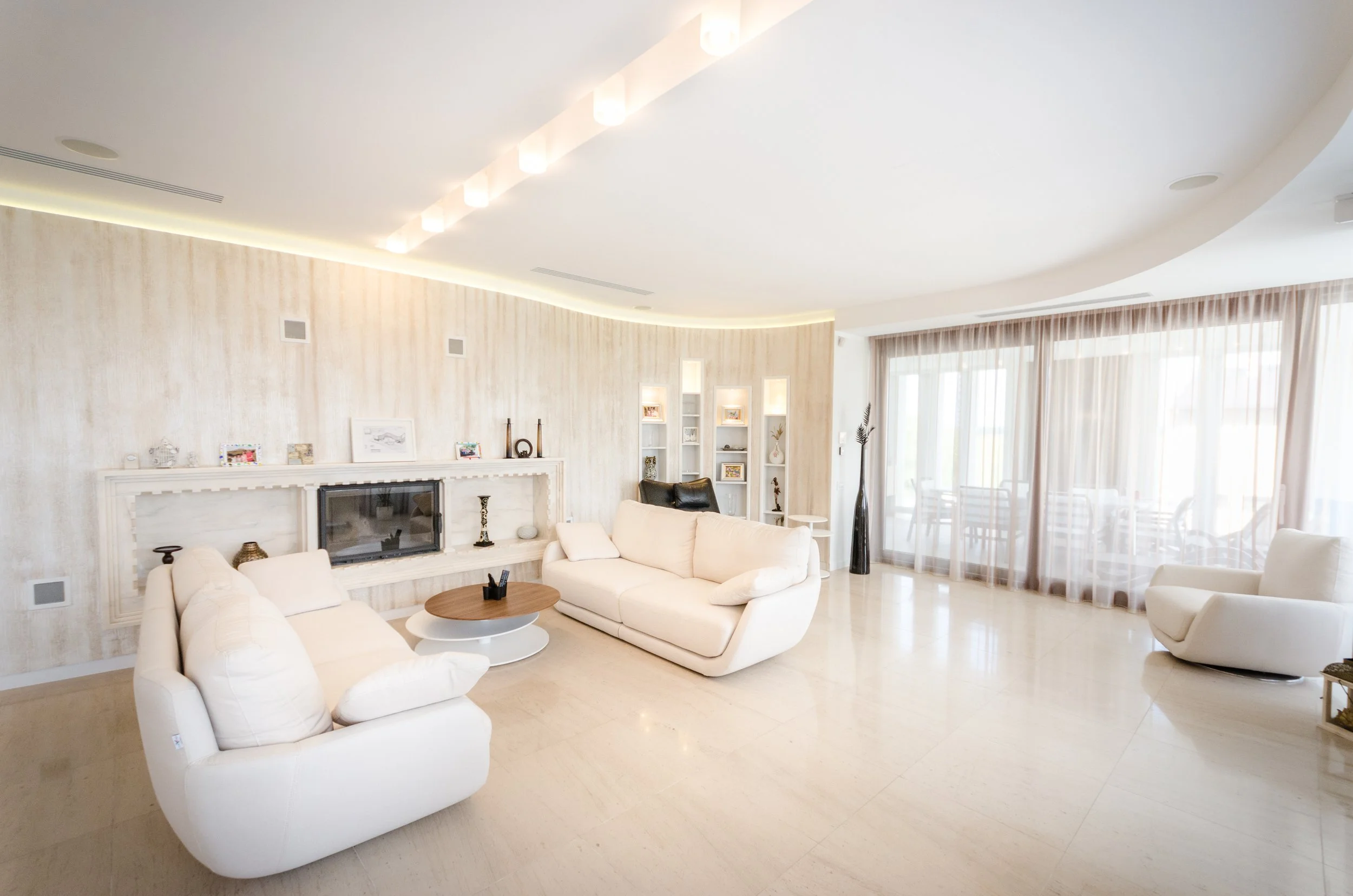 Bright, modern living room with white sofas, a round wooden coffee table, light-colored flooring, large windows with sheer curtains, and built-in wall shelves with decorative items.
