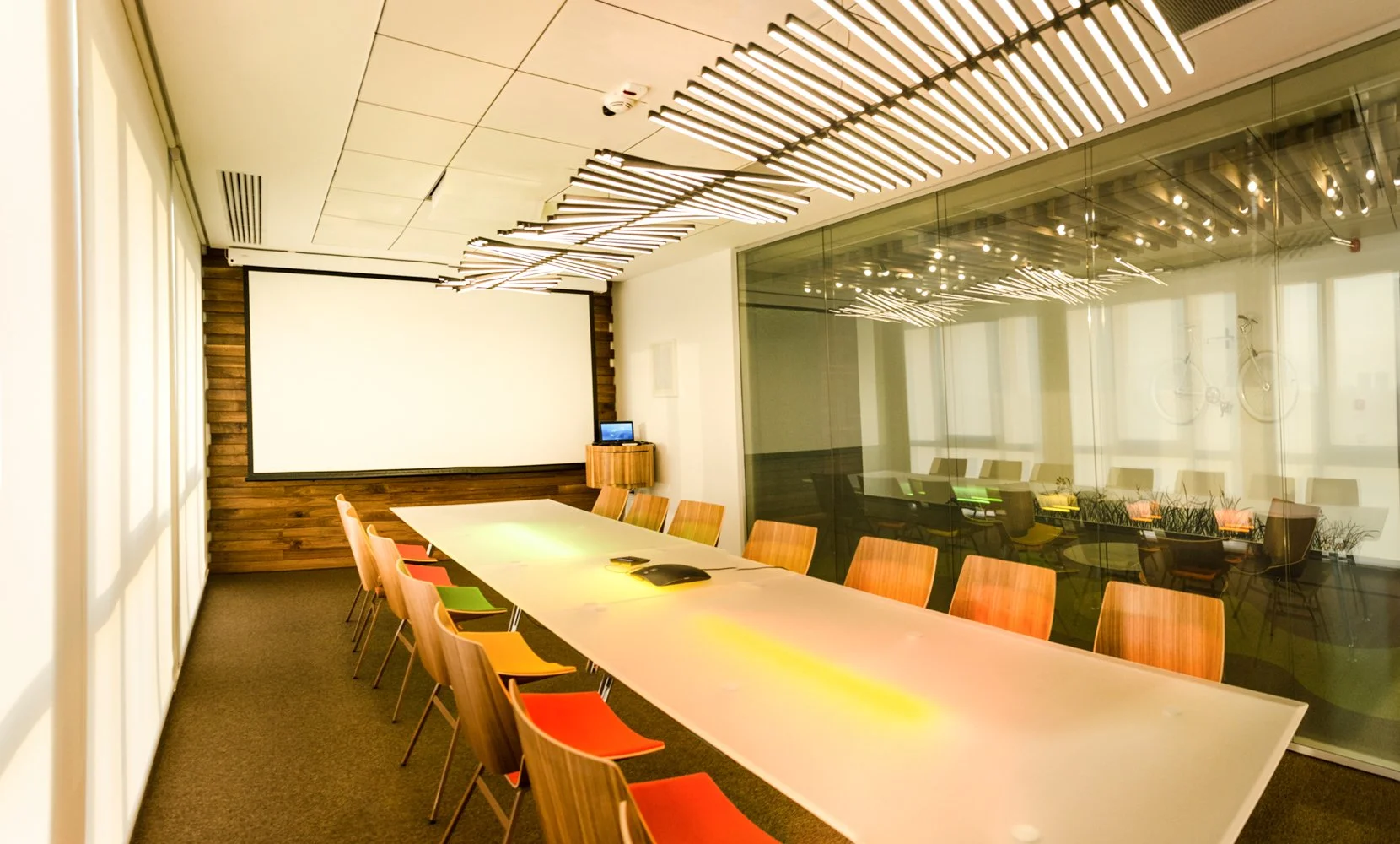 Modern conference room with a long table, colorful chairs, a large projection screen, and contemporary lighting fixtures on the ceiling.