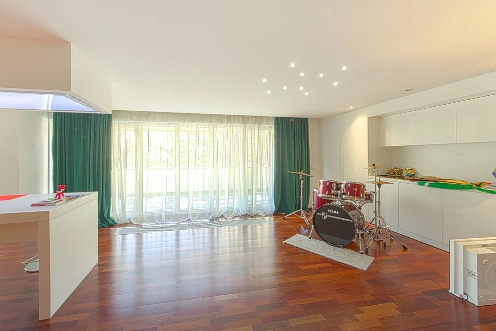 Living room with hardwood floors, large window with sheer white and dark green curtains, and a red drum set with cymbals near the right wall.