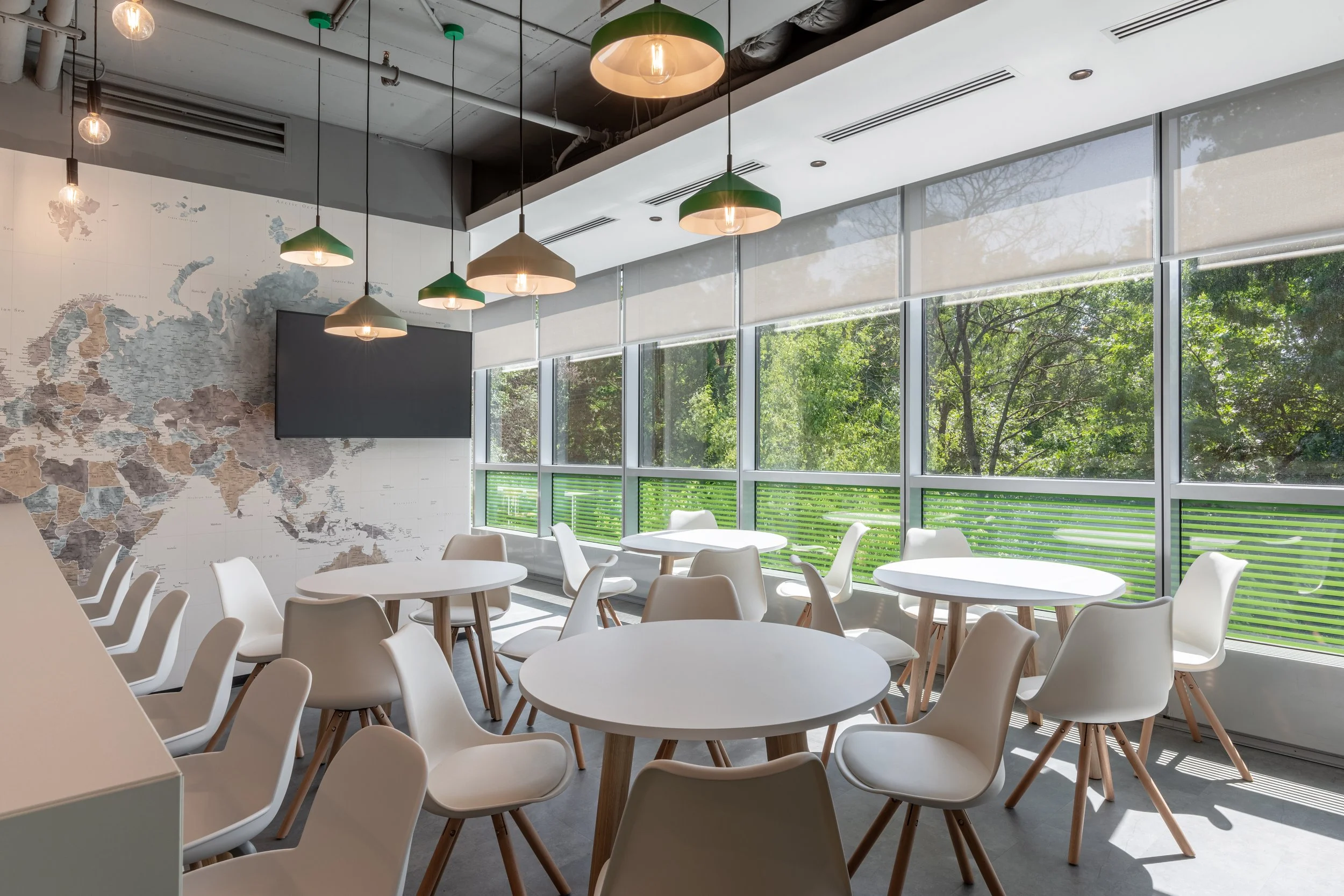 Bright and modern cafe with white round tables and white chairs, large windows with blinds, green and white pendant lights, and a world map on the wall.