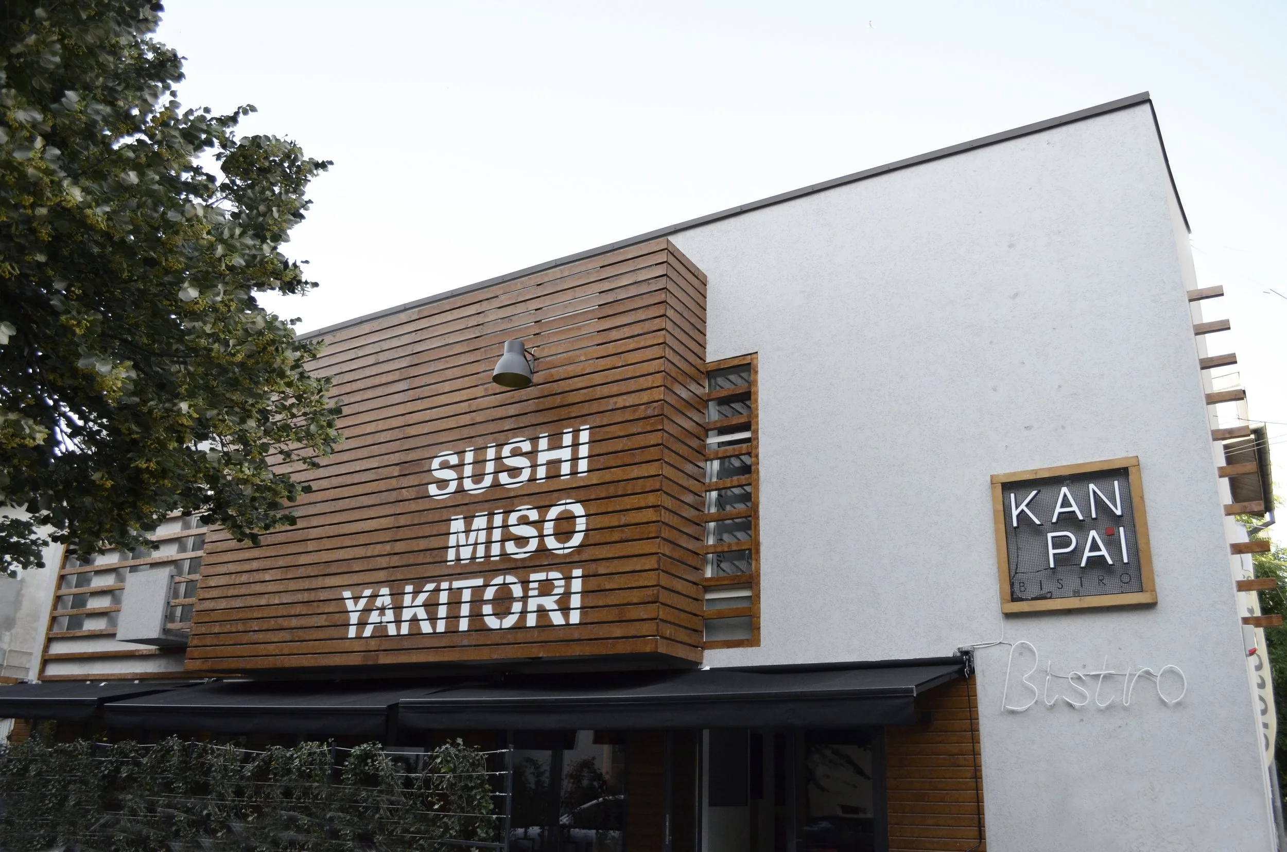 Exterior view of a modern bistro with a white building facade and wooden accents. Large sign on the wall reads "SUSHI MISO YAKITORI" and a smaller sign displays "KAN PAI BISTRO". There are horizontal wooden slats and a black awning.