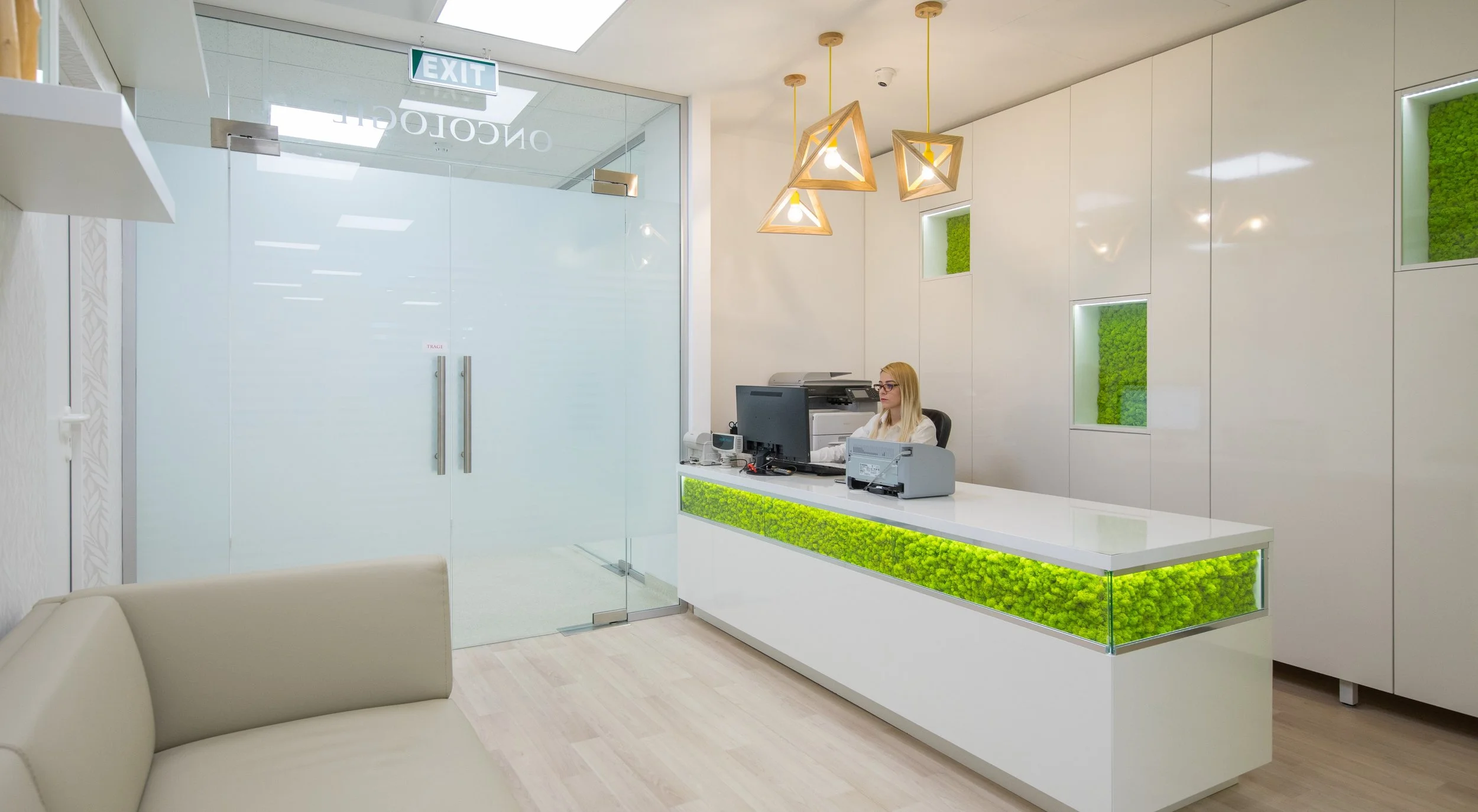 Office reception area with a white counter featuring green moss decor, a woman seated at the desk using a computer, modern hanging light fixtures, glass door, and green moss wall accents.